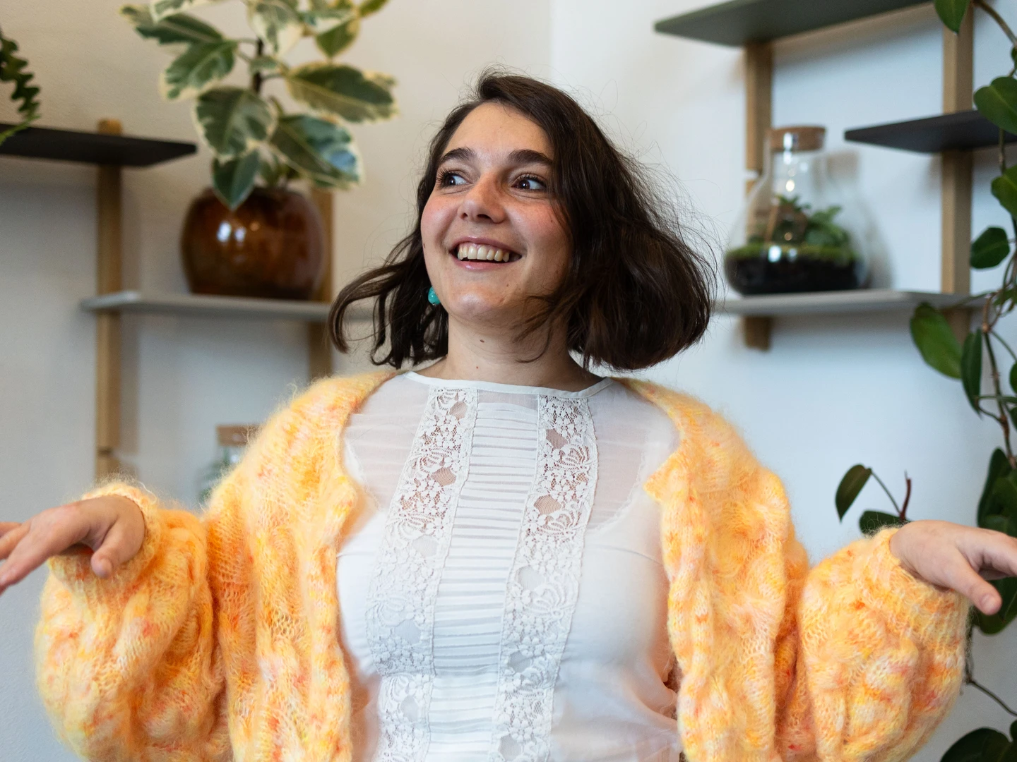 En smilende kvinde med kort hår i en farverig sweater står i et lyst rum med grønne planter og hylder i baggrunden. Hun gestikulerer livligt, hvilket tilføjer en glad og indbydende atmosfære.