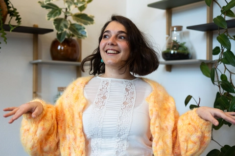 En smilende kvinde med kort hår i en farverig sweater står i et lyst rum med grønne planter og hylder i baggrunden. Hun gestikulerer livligt, hvilket tilføjer en glad og indbydende atmosfære.
