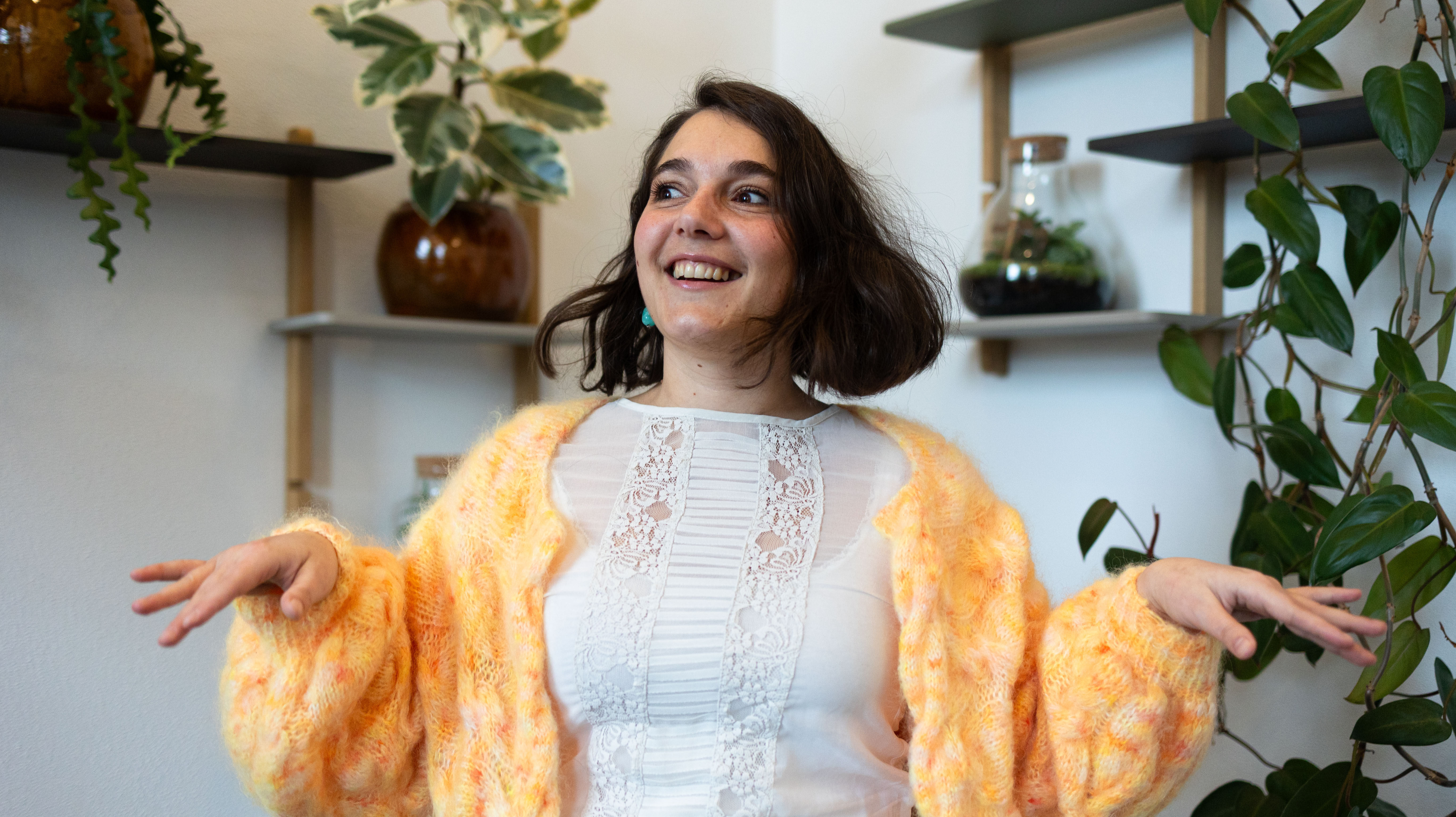 En smilende kvinde med kort hår i en farverig sweater står i et lyst rum med grønne planter og hylder i baggrunden. Hun gestikulerer livligt, hvilket tilføjer en glad og indbydende atmosfære.