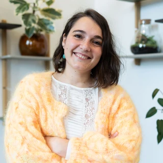 Ung kvinde med kort, bølget hår iført en fluffy, orange cardigan står med armene over kors og smiler, mens hun står foran hylder med planter i et lyst rum.