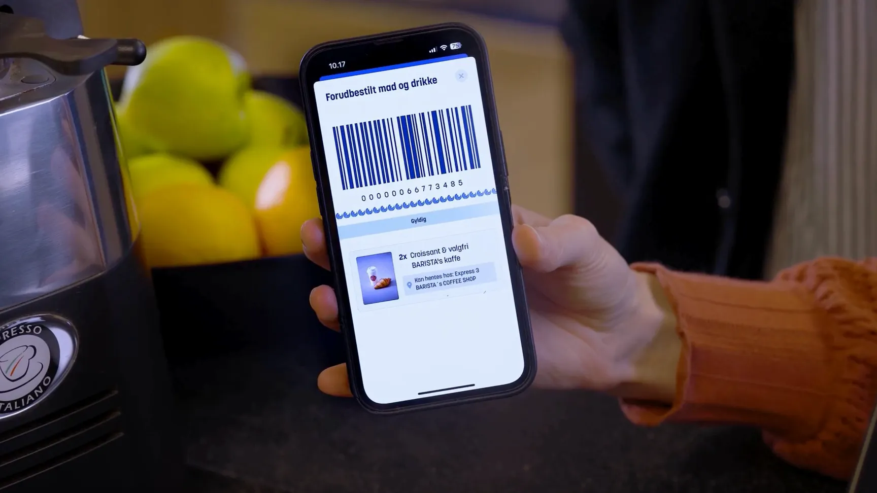 Mobiltelefon viser en forudbestilling af mad og drikke med en stregkode, mens en hånd holder telefonen. I baggrunden ses en skål med æbler og citroner.