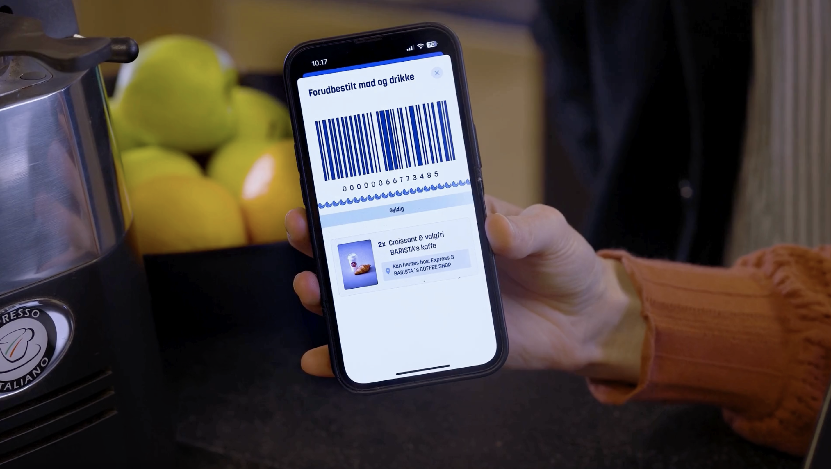Mobiltelefon viser en forudbestilling af mad og drikke med en stregkode, mens en hånd holder telefonen. I baggrunden ses en skål med æbler og citroner.