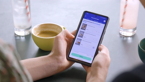 En person, der holder en smartphone med en app til bestilling af mad og drikke, mens der står en kop kaffe og et glas vand på bordet.