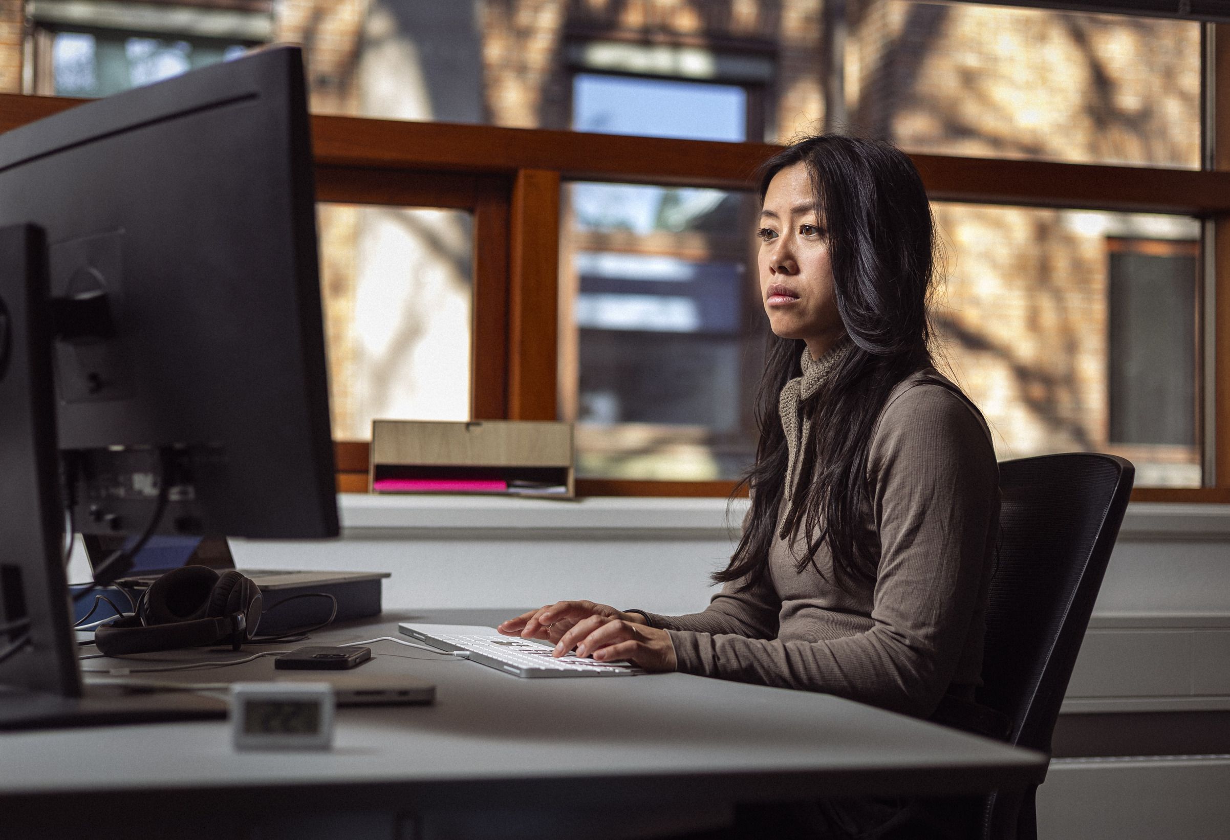 En kvinde sidder ved et skrivebord og arbejder ved en computer, iført en grå trøje og med mørkt, langt hår. Baggrunden viser store vinduer med naturligt lys, der strømmer ind i rummet.