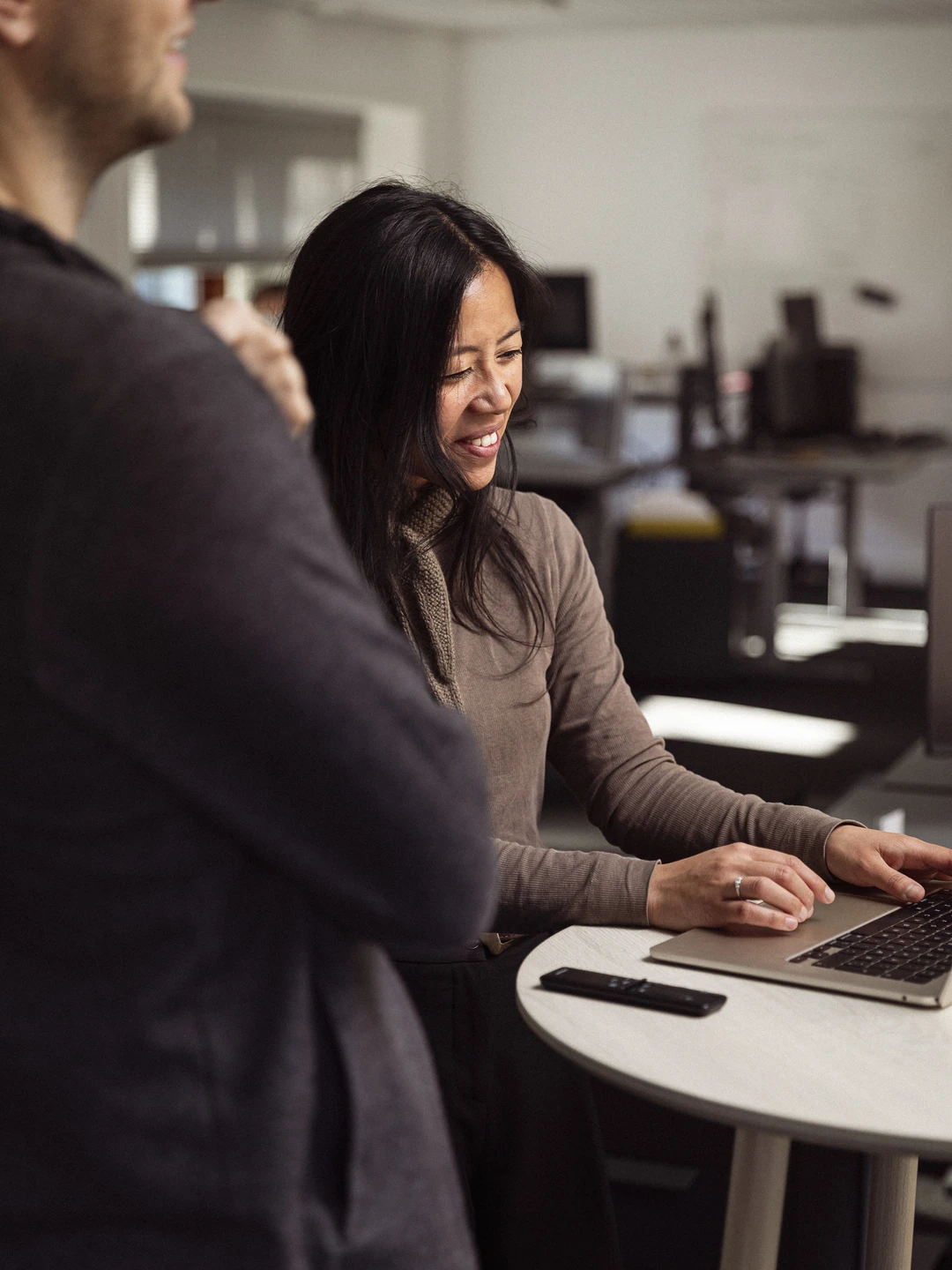 A woman with long hair is smiling while working on a laptop at a small round table in an office setting, with a person partially visible in the foreground.