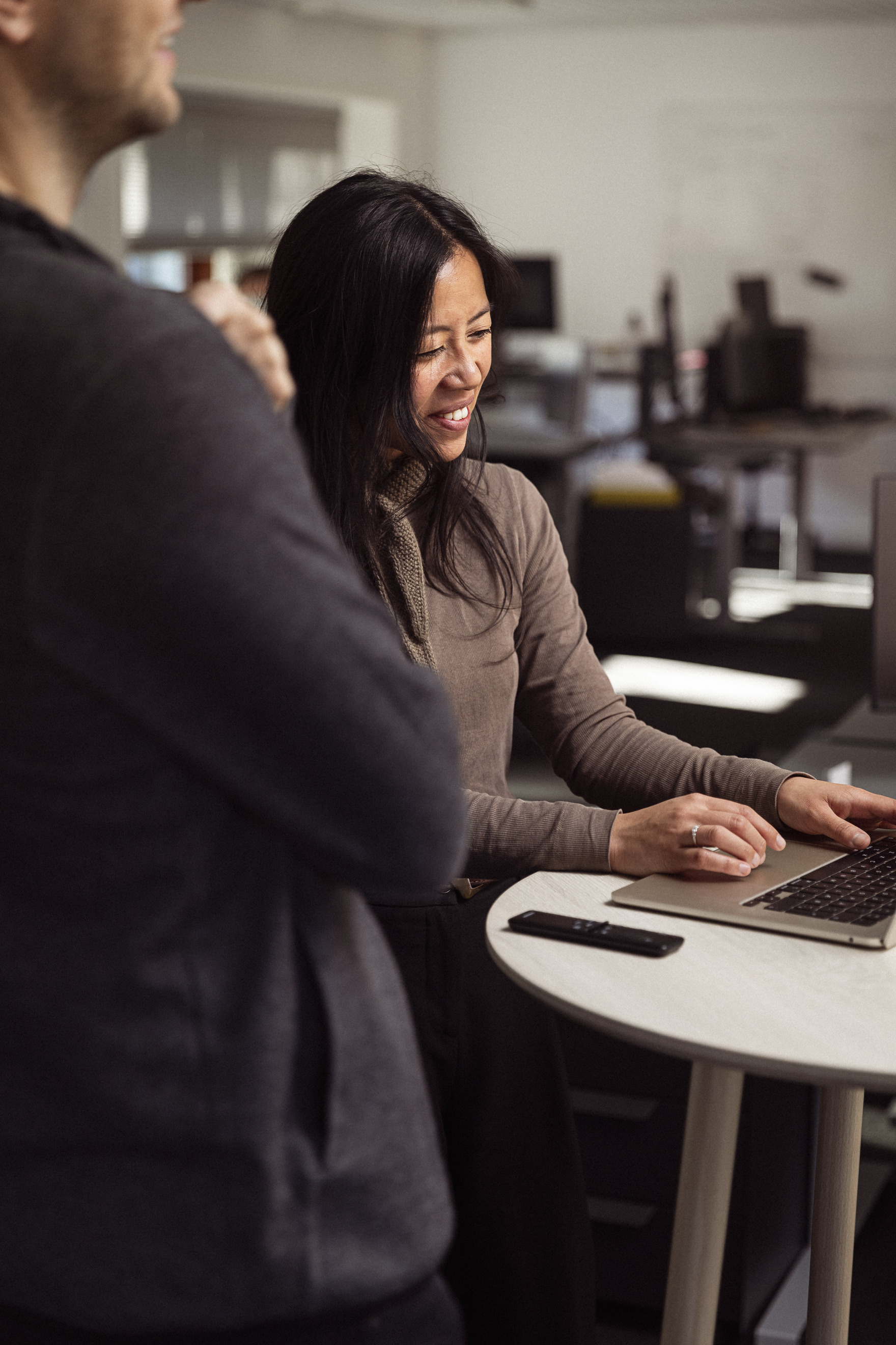 En kvinde med mørkt hår smiler, mens hun arbejder på en bærbar computer, omgivet af et moderne kontormiljø. En mand står ved siden af hende og ser interesseret ud.