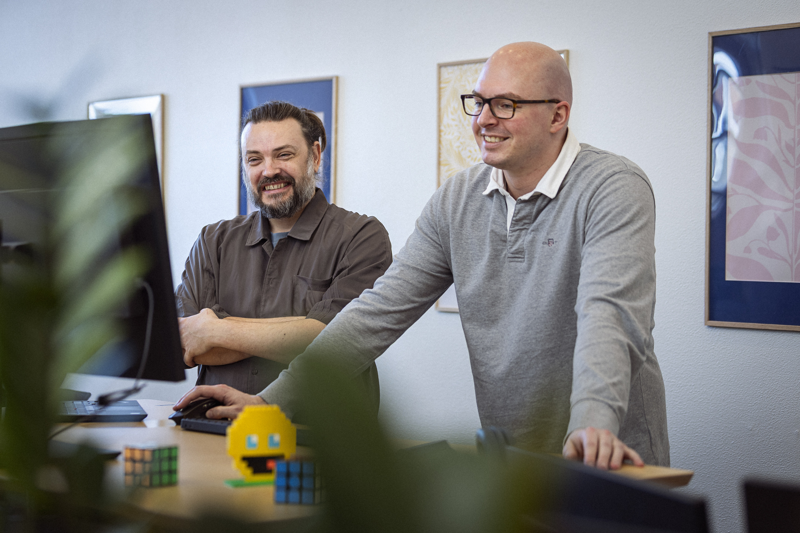 To personer smiler og står sammen ved en computer på et kontor, omgivet af planter og farverige klodser, mens de diskuterer deres arbejde.
