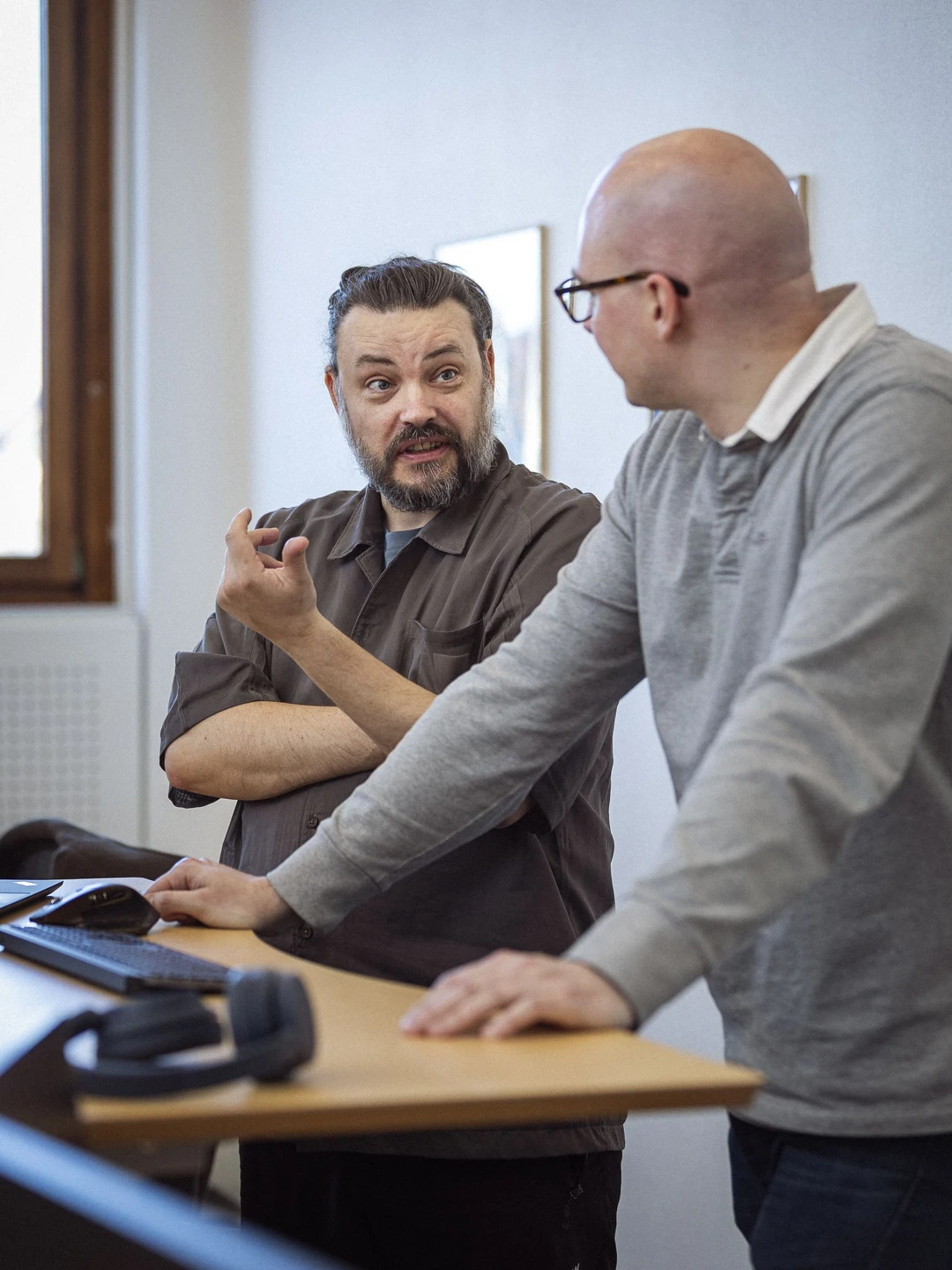 To personer i en diskussion ved et bord med en computer og hovedtelefoner, den første person har skæg og bærer en brun skjorte, mens den anden person er skaldet og iført en grå trøje.