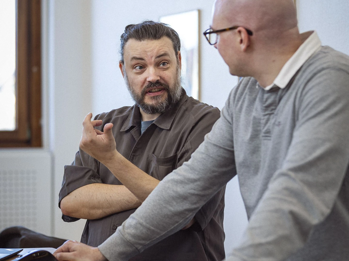 To personer i en diskussion ved et bord med en computer og hovedtelefoner, den første person har skæg og bærer en brun skjorte, mens den anden person er skaldet og iført en grå trøje.