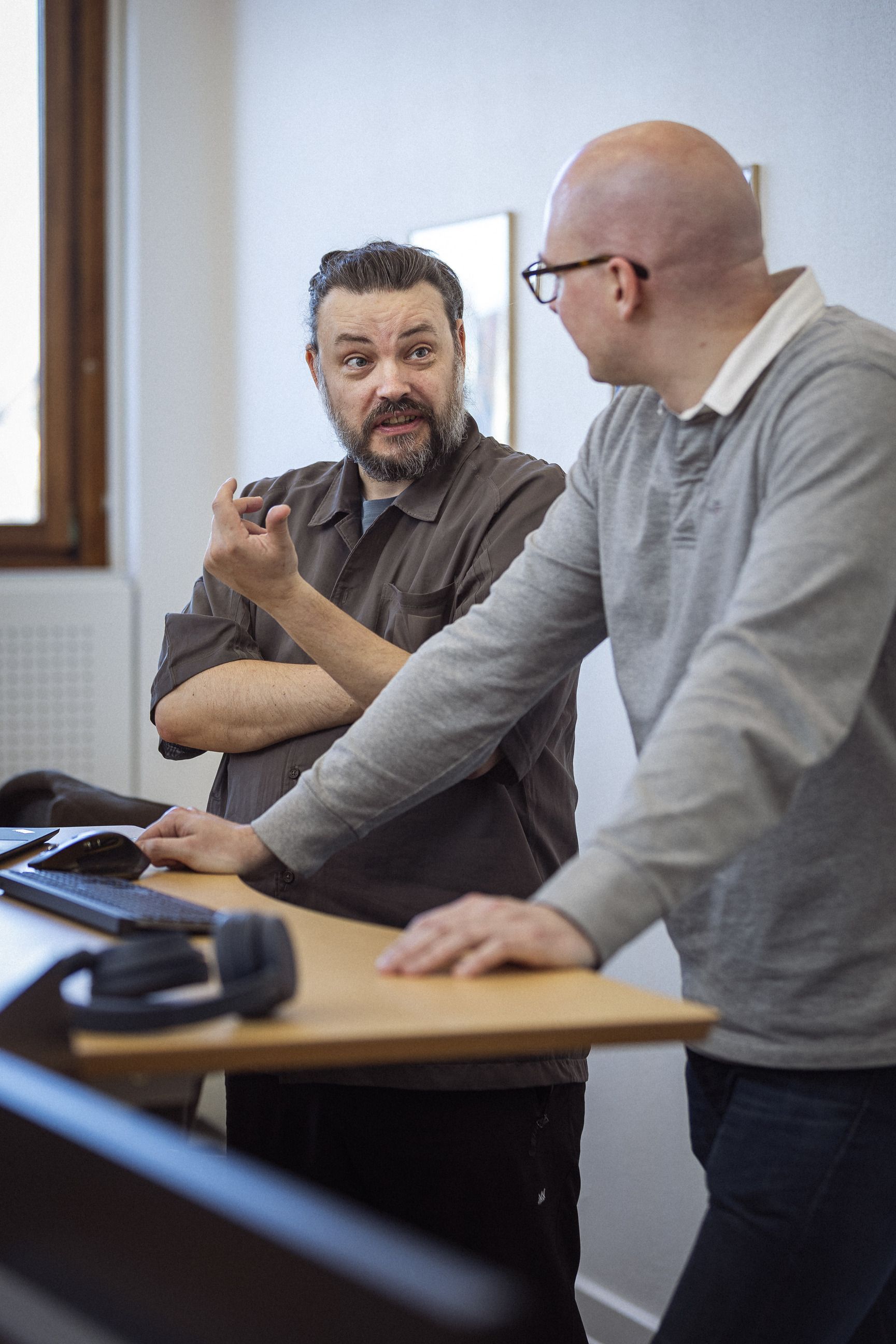 To personer i en diskussion ved et bord med en computer og hovedtelefoner, den første person har skæg og bærer en brun skjorte, mens den anden person er skaldet og iført en grå trøje.