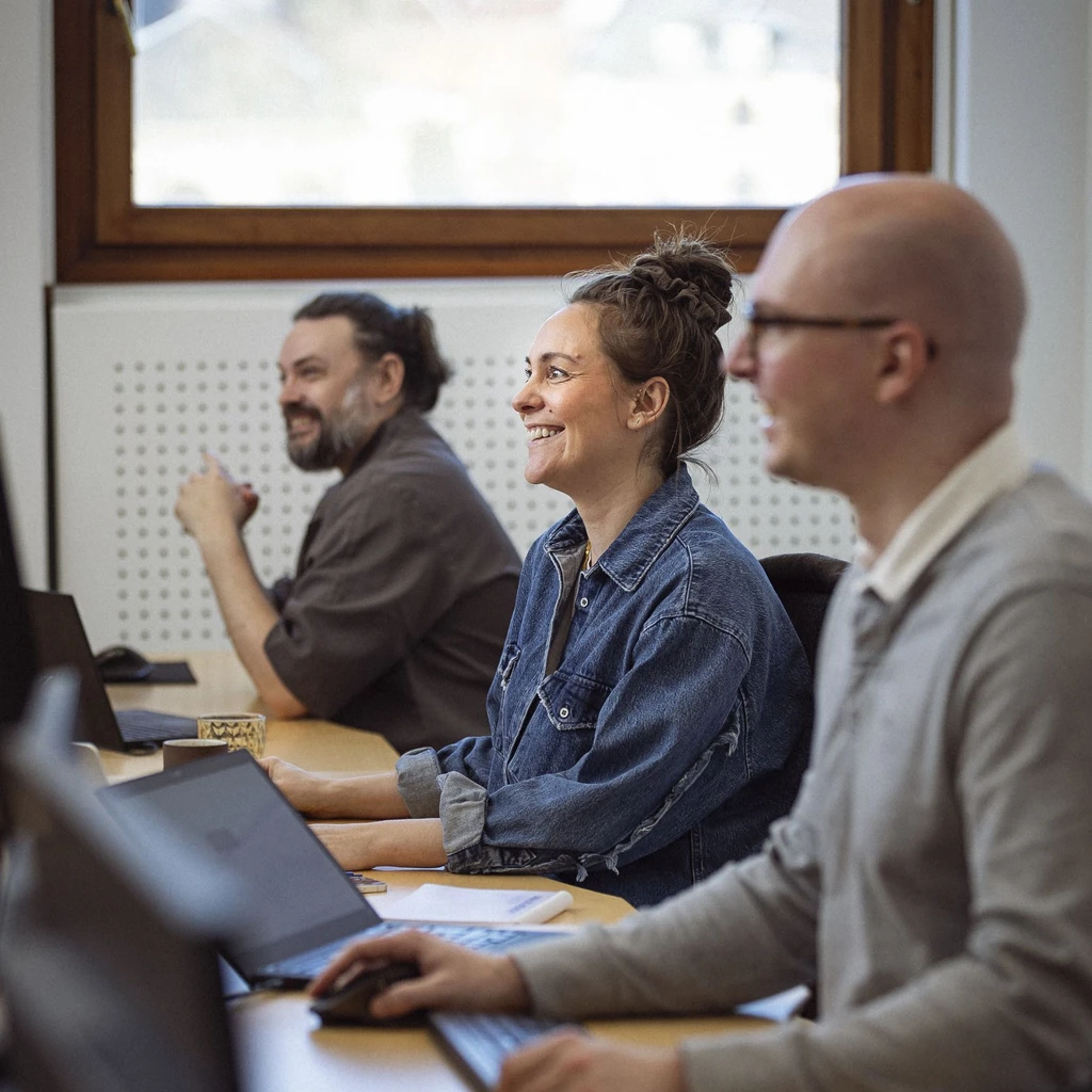 Tre personer, der smiler og arbejder ved computere i et lyst kontor, med fokus på samarbejde og læring.