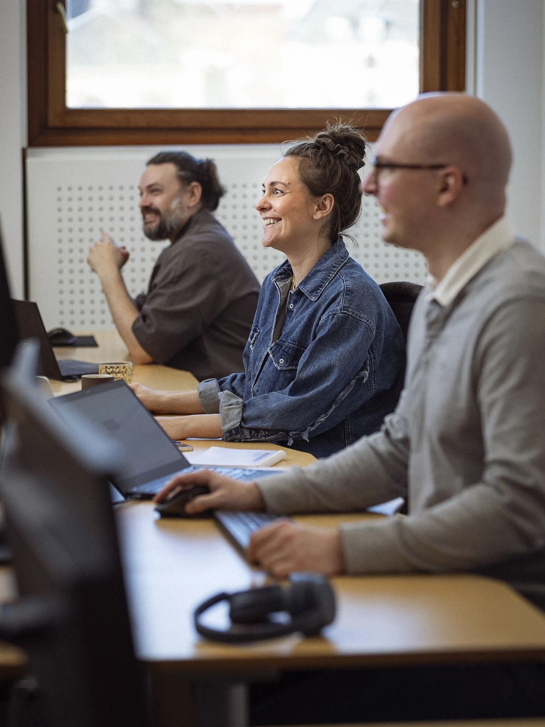 Tre personer, der smiler og arbejder ved computere i et lyst kontor, med fokus på samarbejde og læring.