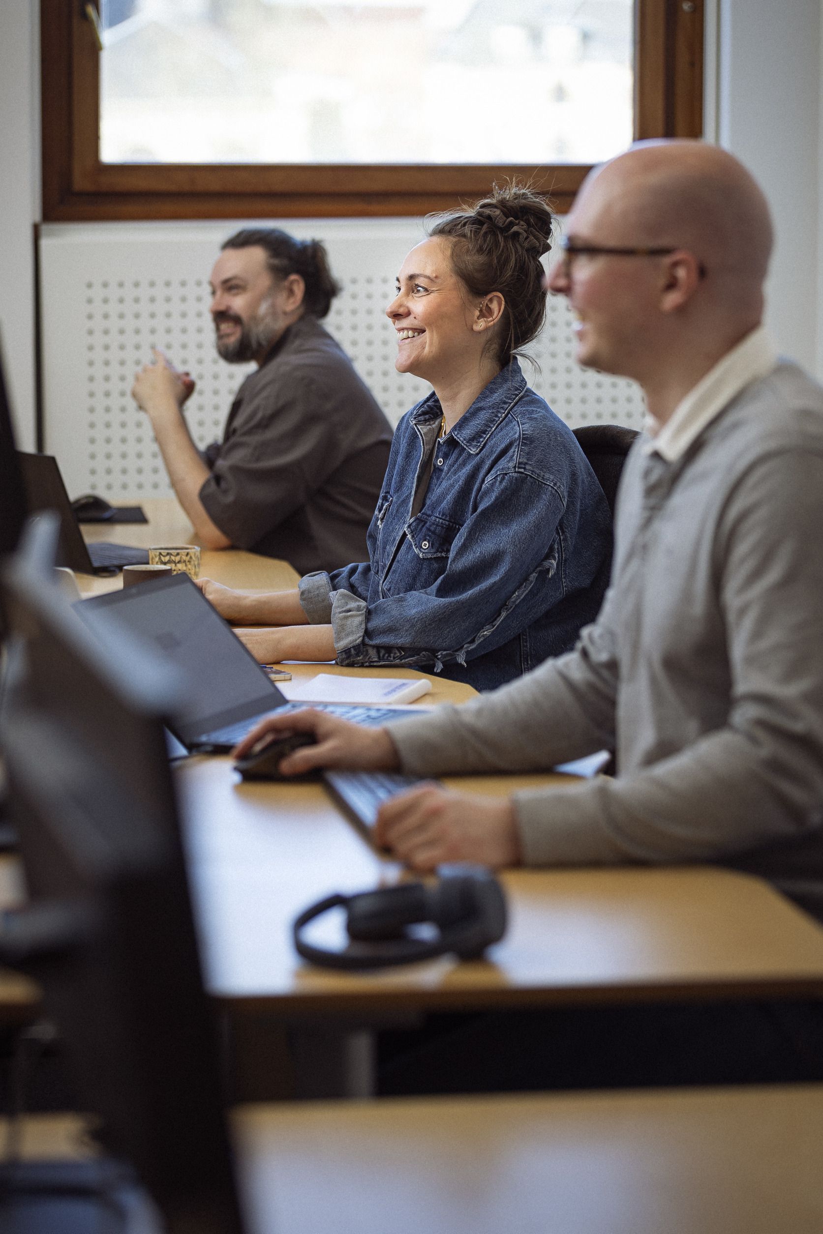 Tre personer, der smiler og arbejder ved computere i et lyst kontor, med fokus på samarbejde og læring.