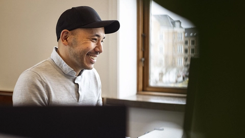 Smiling man in a gray sweater and black cap, sitting at a desk with a computer, with a window and buildings visible in the background.