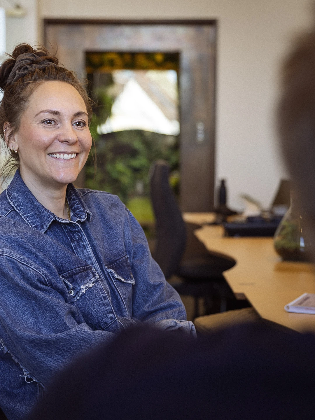 Ung kvinde med smil, iført denimjakke, sidder ved et bord i et lyst kontormiljø, mens hun samtaler med en person udenfor billedet.