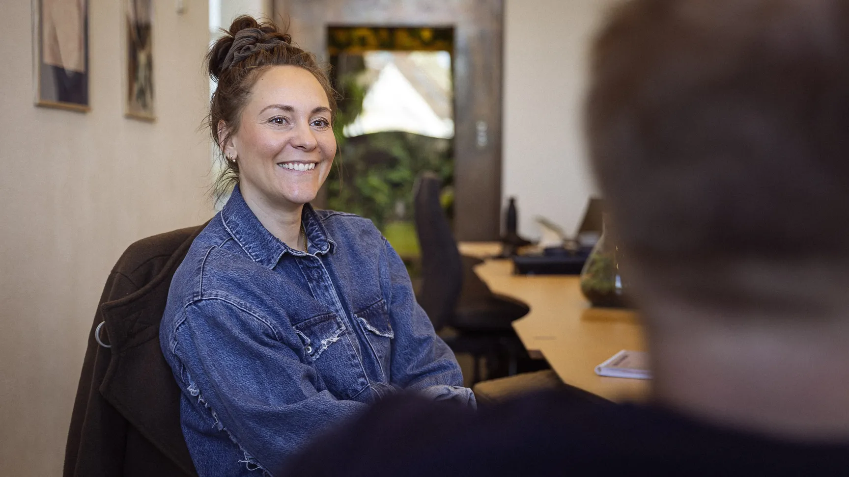 Ung kvinde med smil, iført denimjakke, sidder ved et bord i et lyst kontormiljø, mens hun samtaler med en person udenfor billedet.