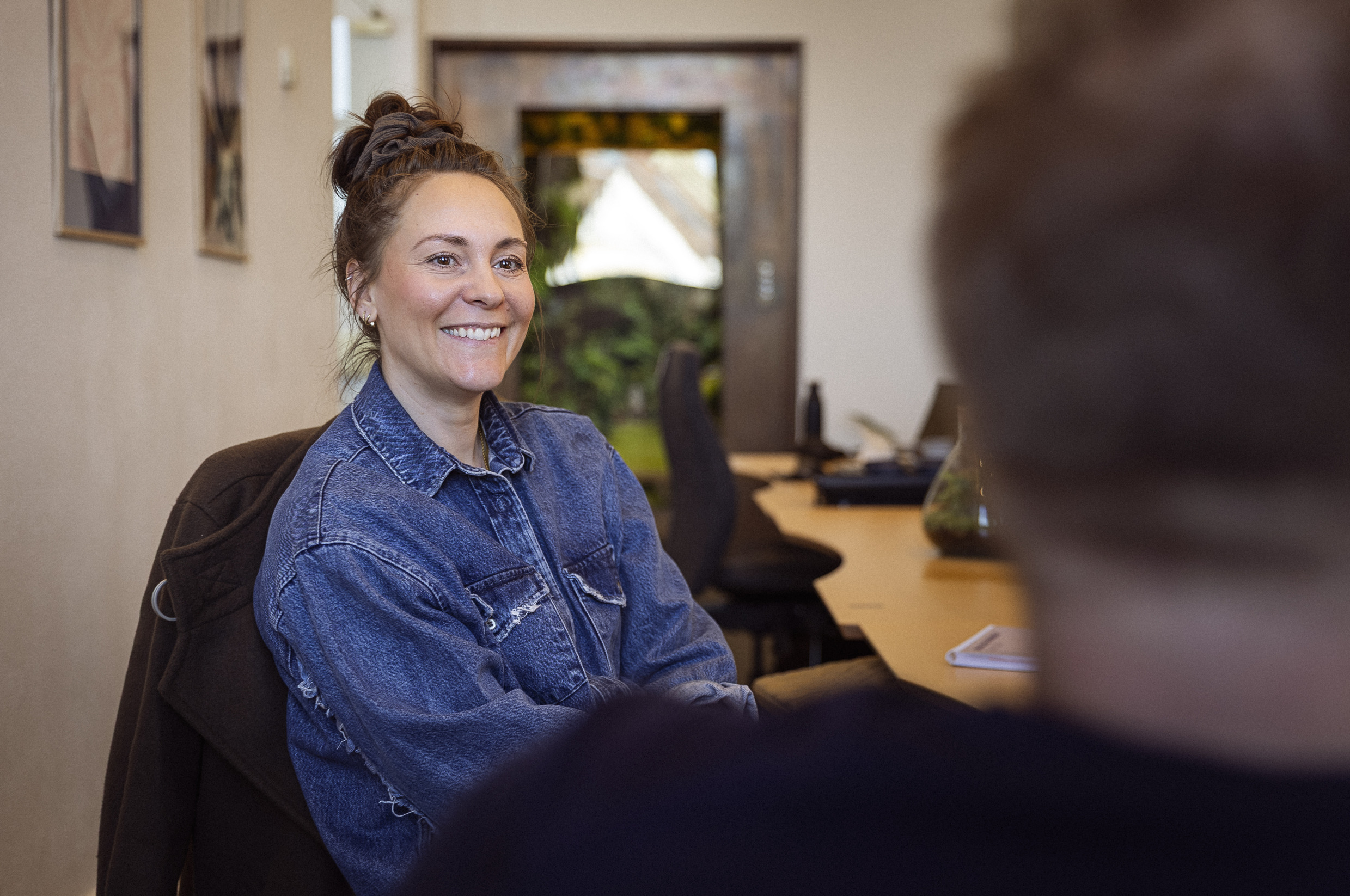 Ung kvinde med smil, iført denimjakke, sidder ved et bord i et lyst kontormiljø, mens hun samtaler med en person udenfor billedet.