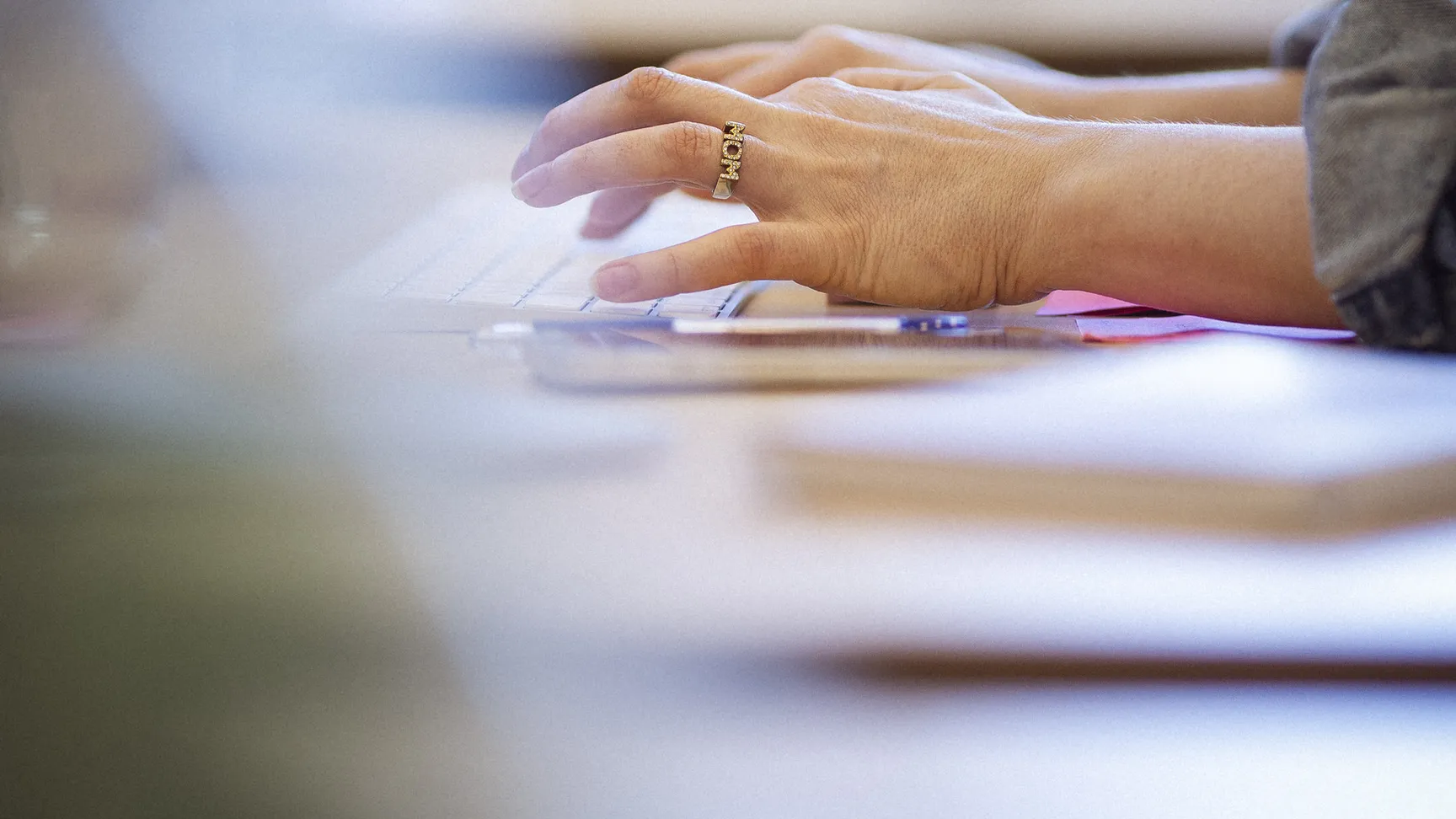 Hænder, der taster på et tastatur, med fokus på en ring på den venstre hånd, i en kontorindstilling med notater i baggrunden.