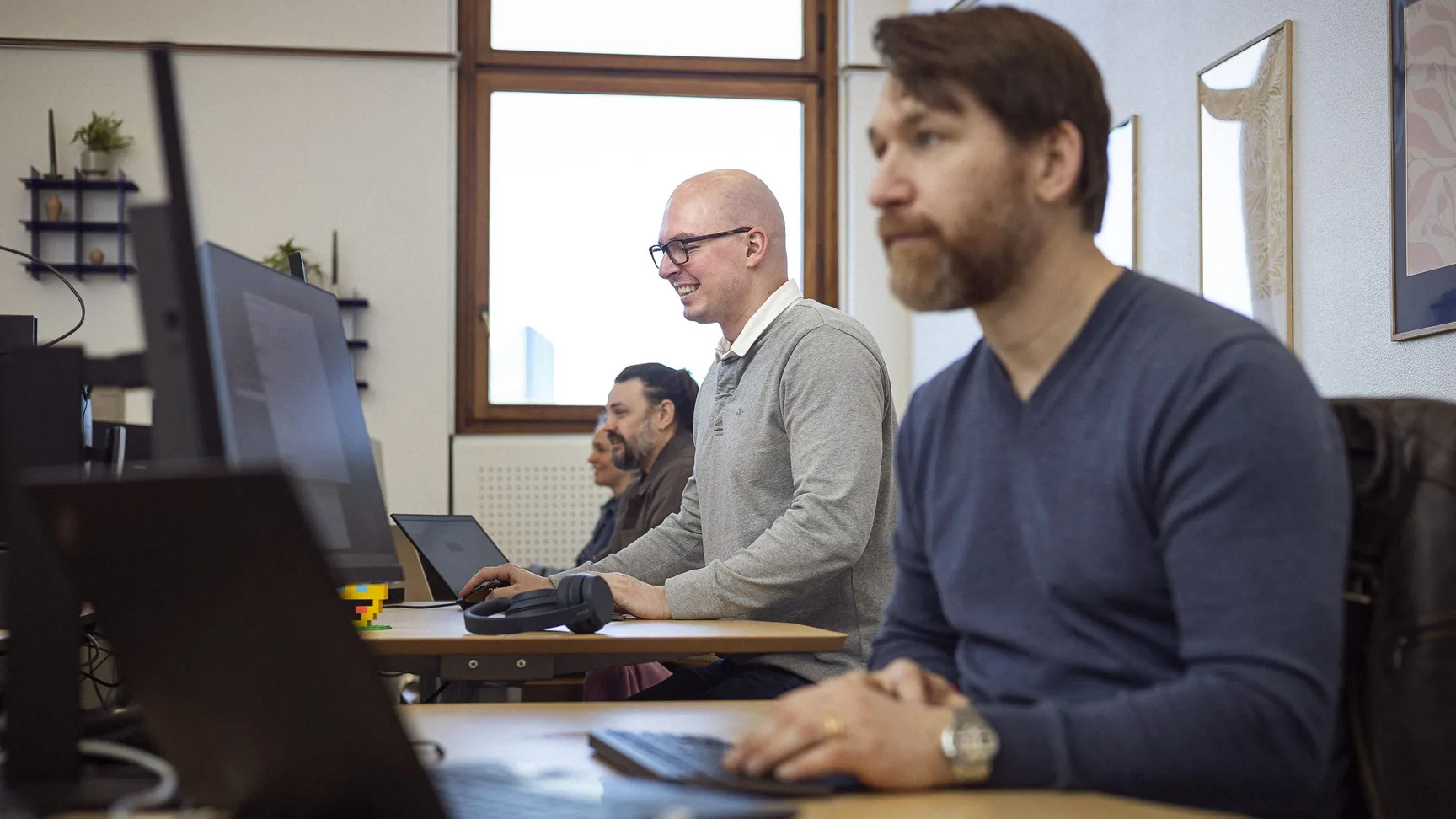 Tre personer arbejder koncentreret ved computere i et lyst kontormiljø med træborde og vinduer, der giver naturligt lys.