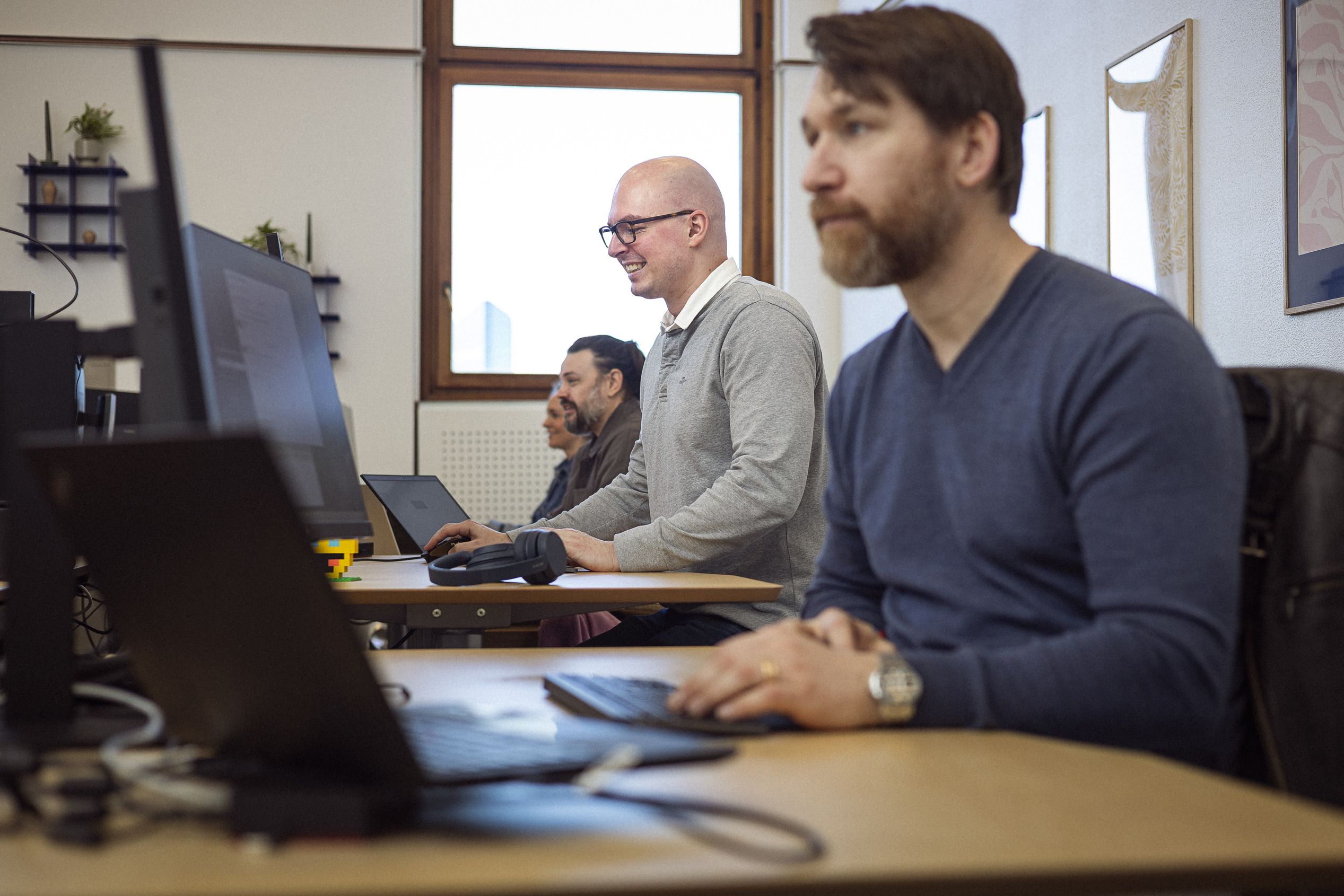 Tre personer arbejder koncentreret ved computere i et lyst kontormiljø med træborde og vinduer, der giver naturligt lys.