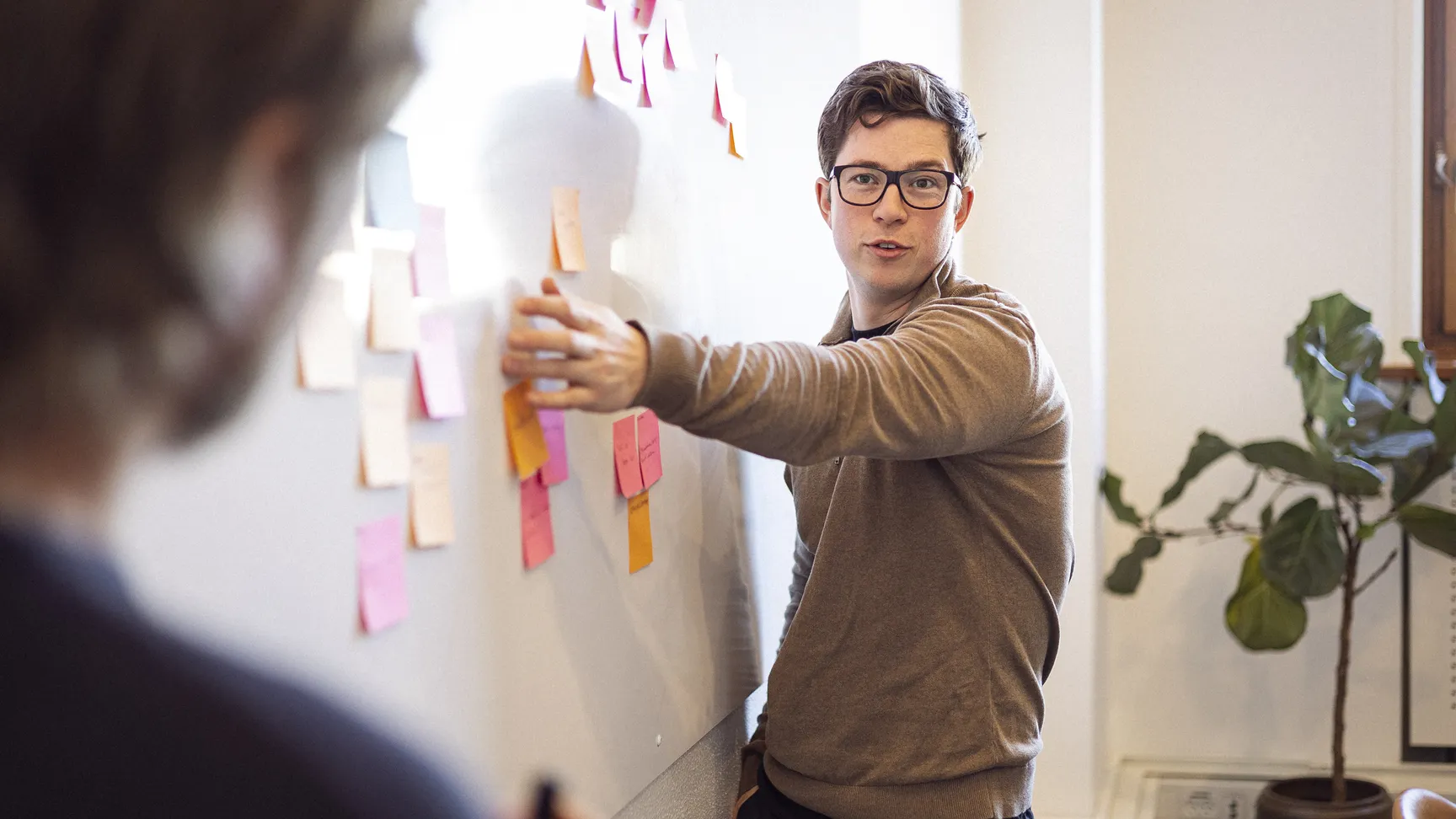 En mand med briller præsenterer idéer ved en opslagstavle dækket med farvede post-it sedler, mens en anden person observerer.