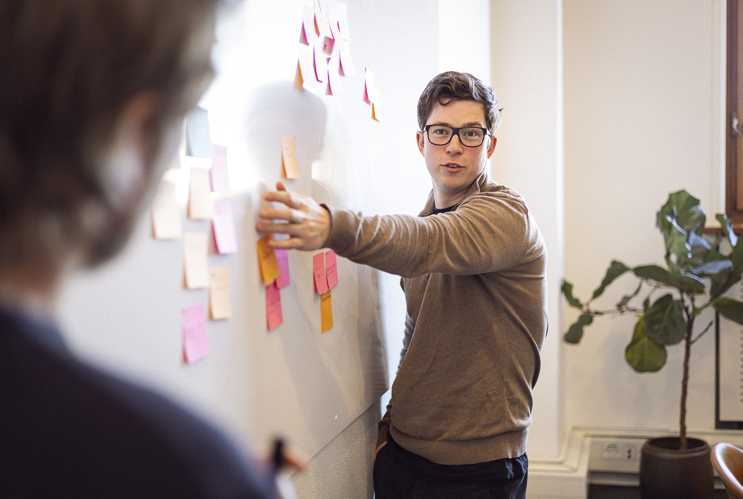 En mand med briller præsenterer idéer ved en opslagstavle dækket med farvede post-it sedler, mens en anden person observerer.