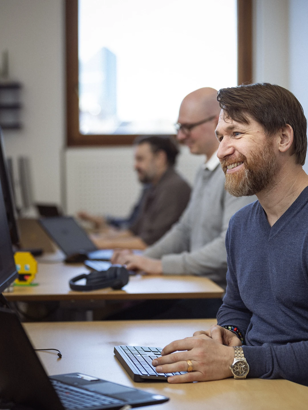 Smilende mand arbejder ved computer. Hans to kollegaer arbejder også i baggrunden.