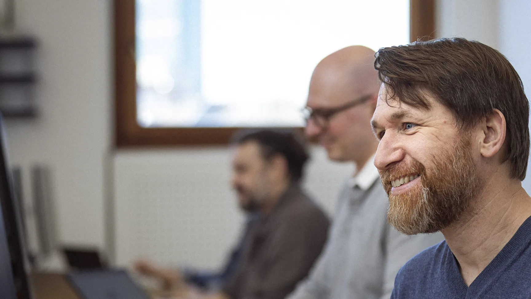 Smilende mand arbejder ved computer. Hans to kollegaer arbejder også i baggrunden.