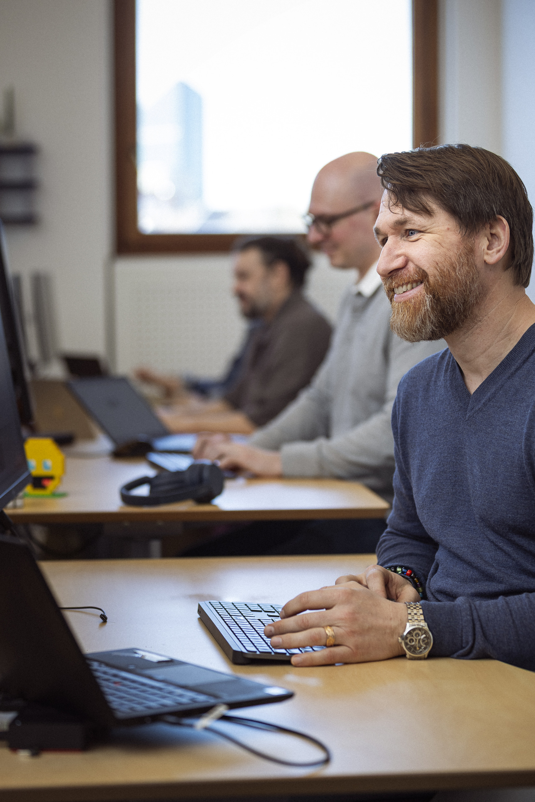 Smilende mand arbejder ved computer. Hans to kollegaer arbejder også i baggrunden.
