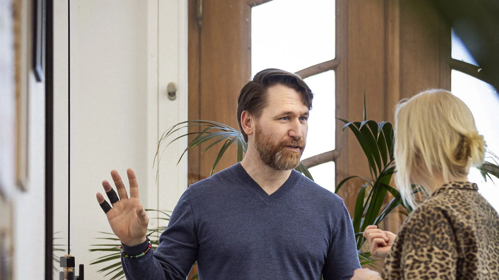 To mænd i samtale i et lyst rum med planter, den ene mand bærer en blå trøje og jeans, og den anden kvinde har en leopardmønstret bluse og jeans, diskussionen ser ud til at være engagerende.