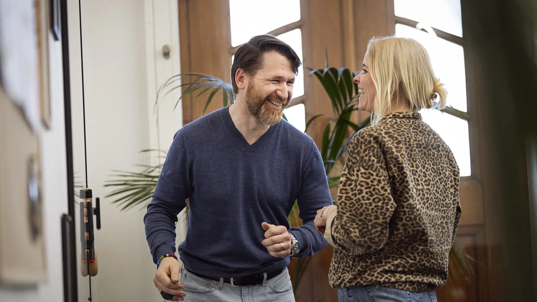 To mennesker griner og interagerer i et lyst rum med planter i baggrunden, iført afslappet tøj.