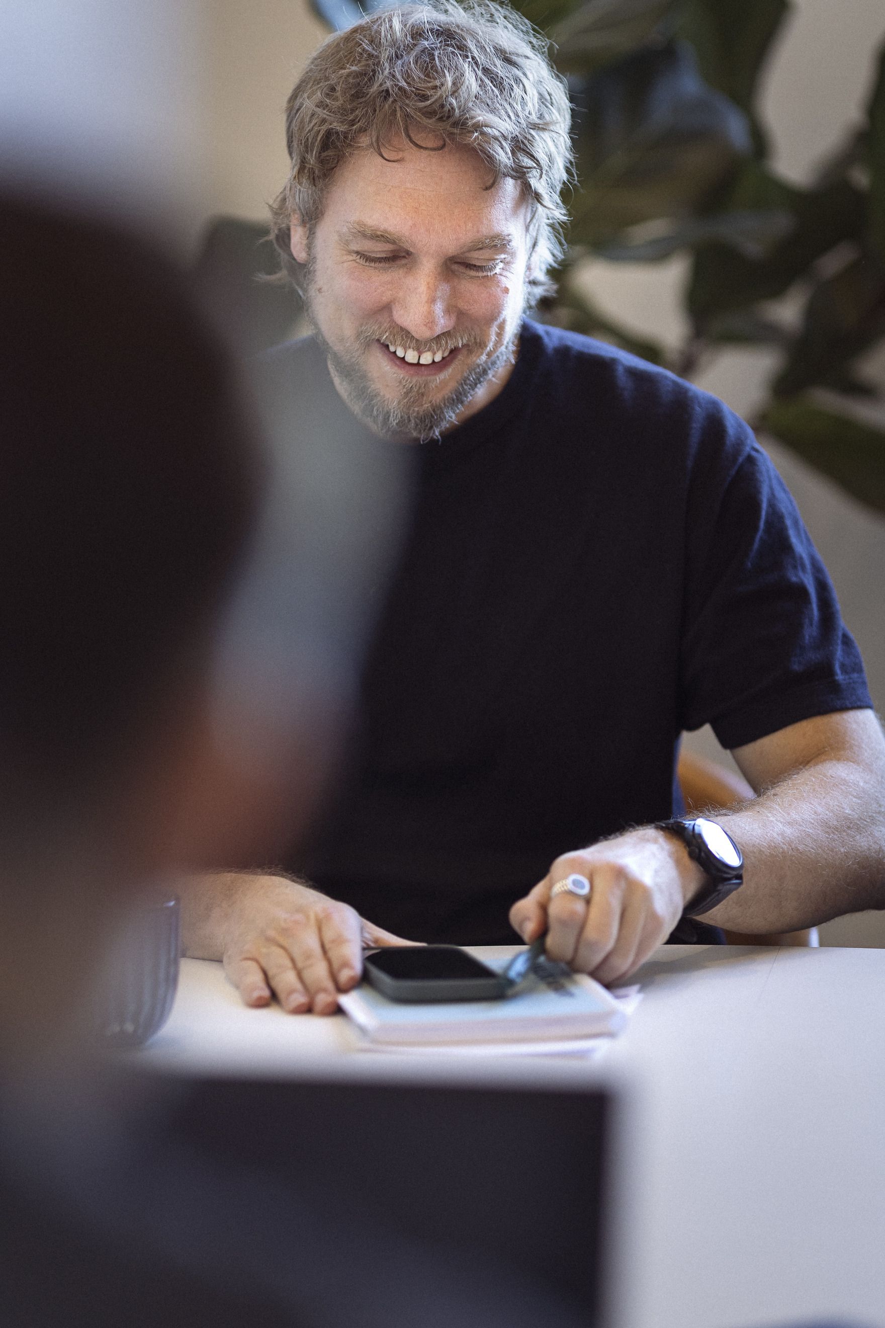 En smilende mand med krøllet hår sidder ved et bord, holder en smartphone og bladrer i en blok med papir, mens han ser glad ud.