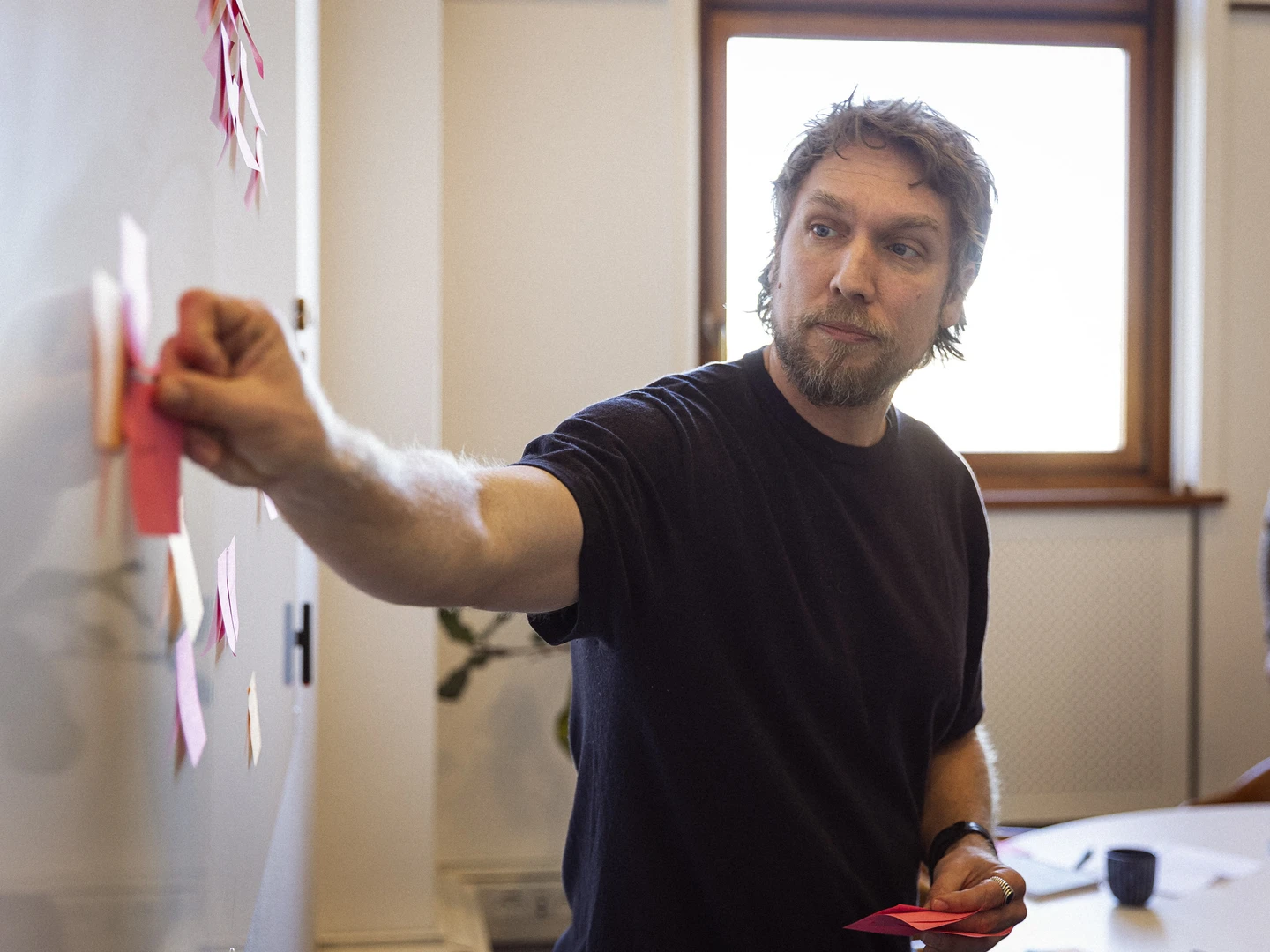 Mand med skæg peger på en tavle med farvede post-its under et møde i et lyst rum med vindue.