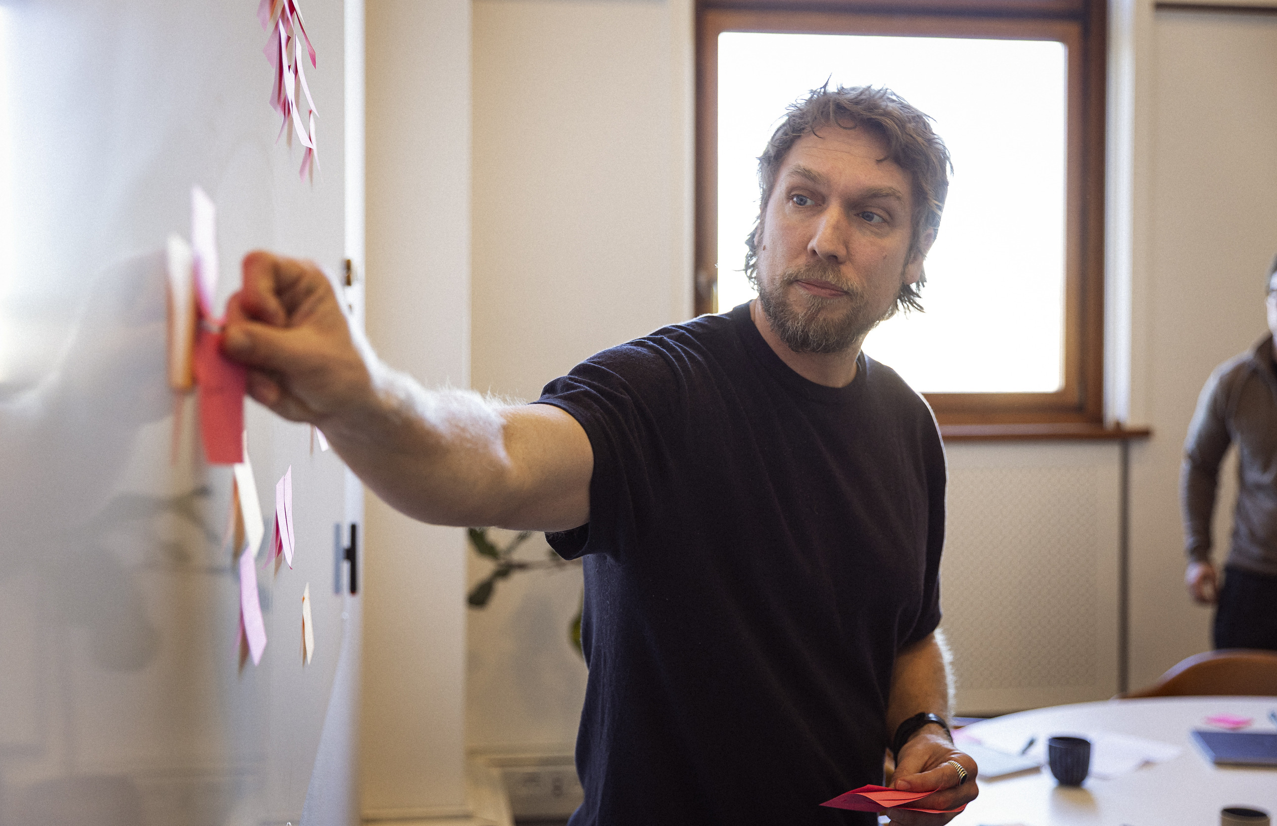 Mand med skæg peger på en tavle med farvede post-its under et møde i et lyst rum med vindue.