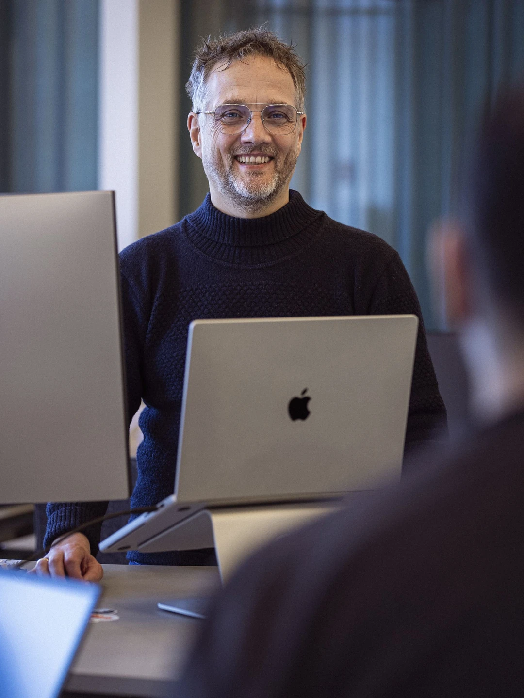 En smilende mand i en mørk striktrøje står foran en laptop på et kontor, med et professionelt og indbydende miljø.