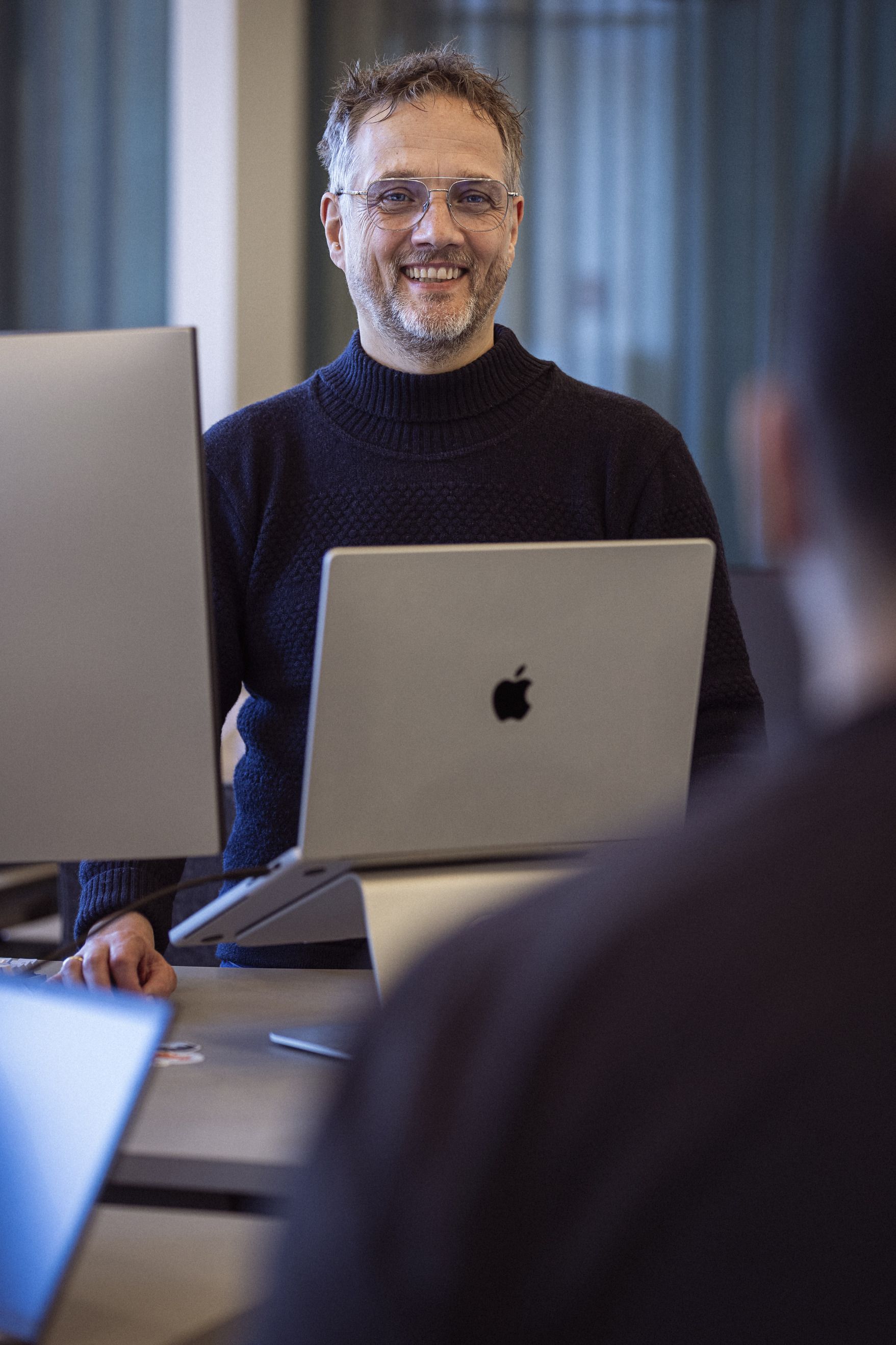 En smilende mand i en mørk striktrøje står foran en laptop på et kontor, med et professionelt og indbydende miljø.