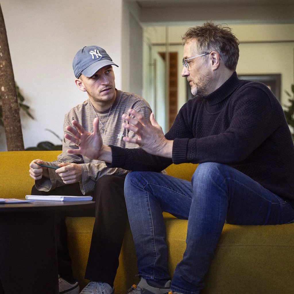 To personer, der sidder på en gul sofa, engageret i en samtale, mens de gestikulerer med hænderne, med planter i baggrunden og et bord med bøger og notater foran dem.