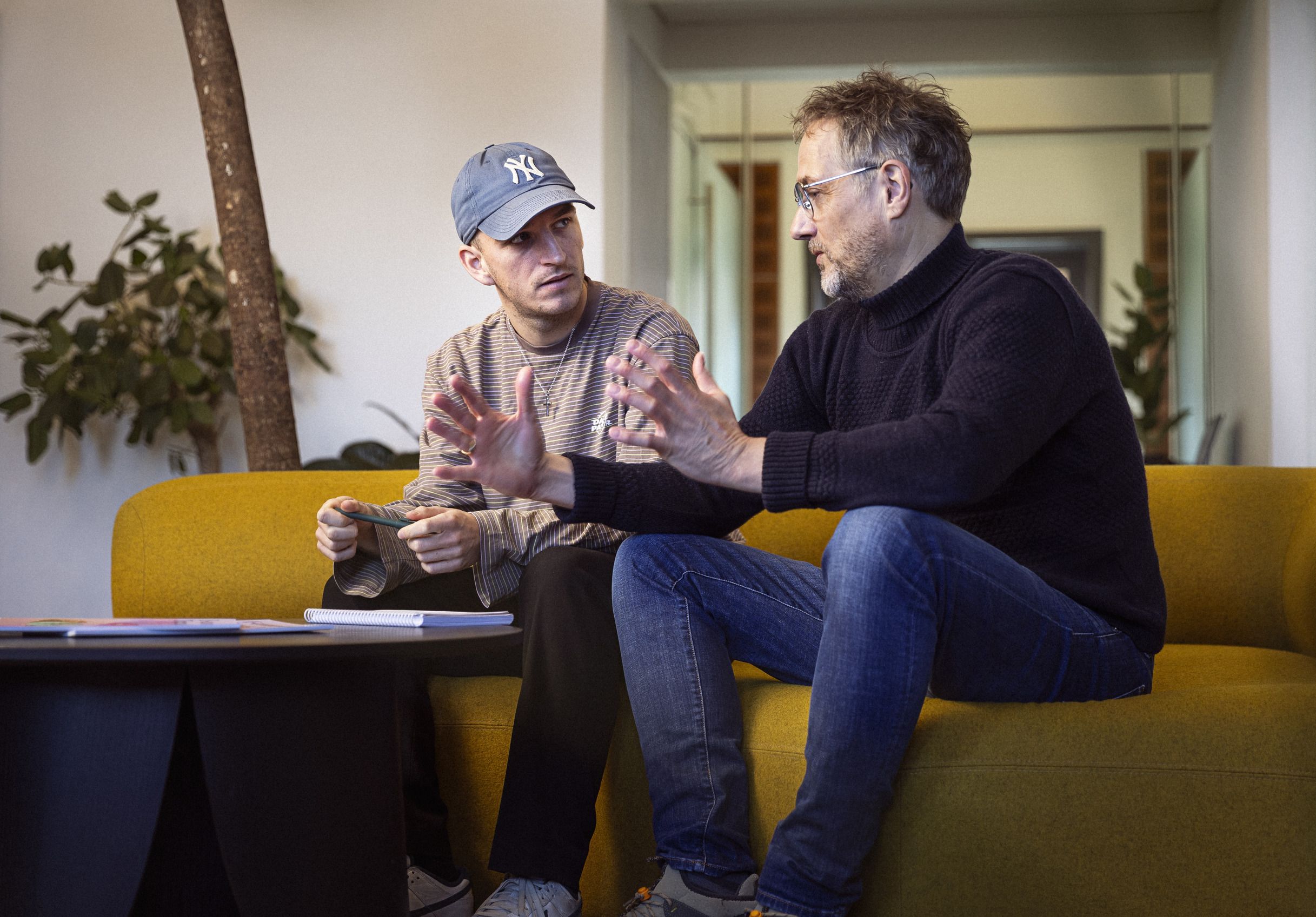 To personer, der sidder på en gul sofa, engageret i en samtale, mens de gestikulerer med hænderne, med planter i baggrunden og et bord med bøger og notater foran dem.