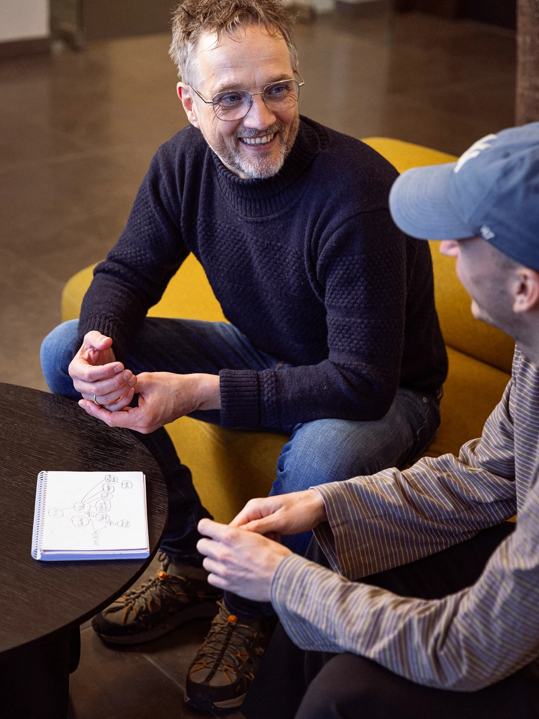 To personer sidder og taler sammen i et moderne rum. Den ene person har gråligt hår og briller, iført en mørk sweater, og smiler mens han holder en rød genstand. Den anden person bærer en blå cap og en stribet skjorte, og de to ser ud til at diskutere noget på en notesblok med skitser. En gul sofa og et mørkt bord kan ses i baggrunden.