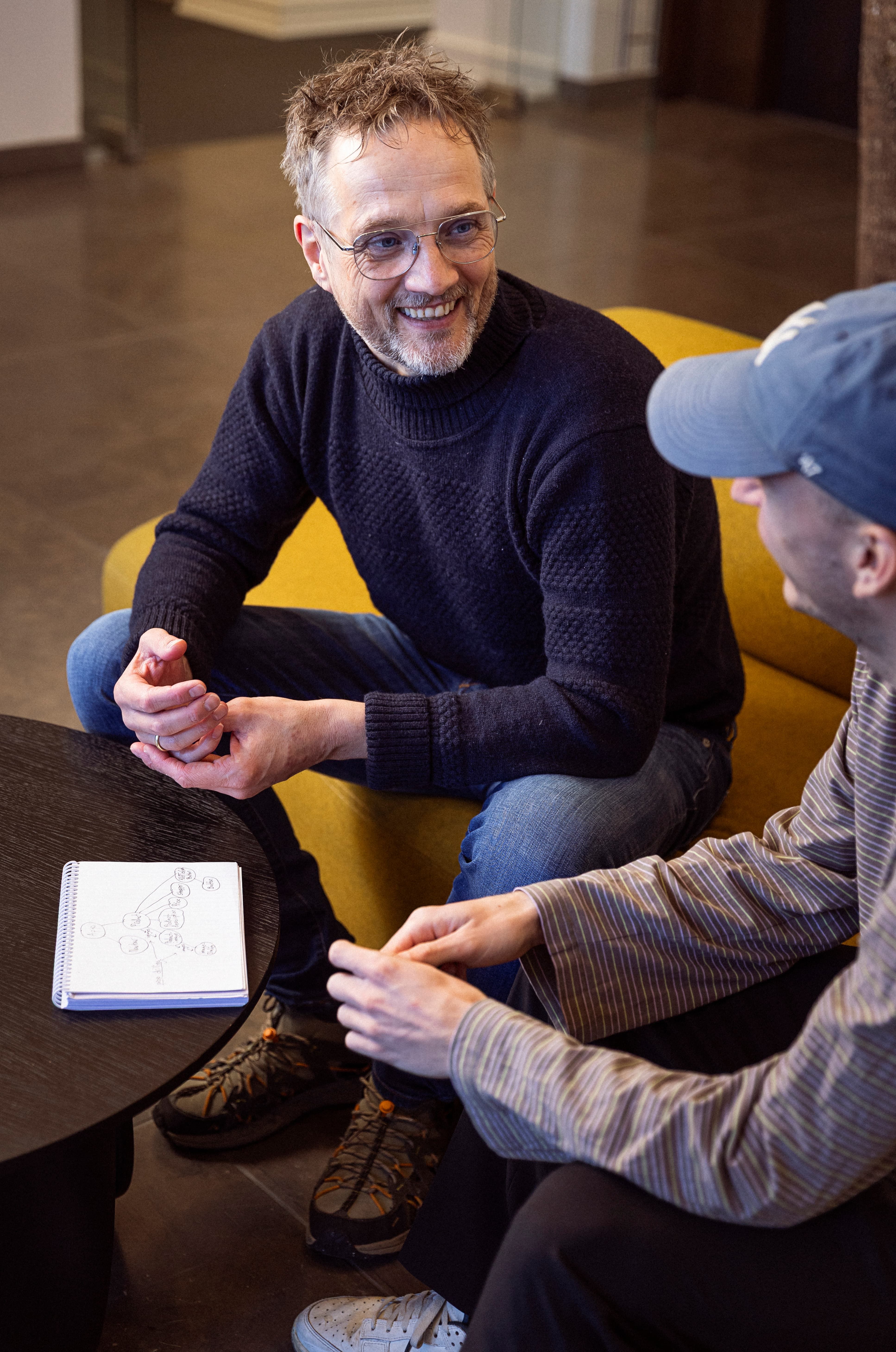 To personer sidder og taler sammen i et moderne rum. Den ene person har gråligt hår og briller, iført en mørk sweater, og smiler mens han holder en rød genstand. Den anden person bærer en blå cap og en stribet skjorte, og de to ser ud til at diskutere noget på en notesblok med skitser. En gul sofa og et mørkt bord kan ses i baggrunden.