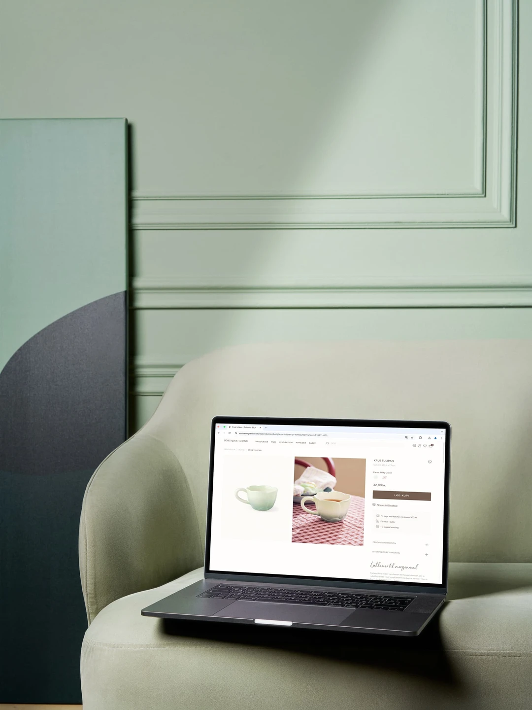 Billede af en bærbar computer, der viser et produktbillede af en keramisk kande på en rødternet dug, placeret på en grøn sofa med en dekorativ vægplade i baggrunden.