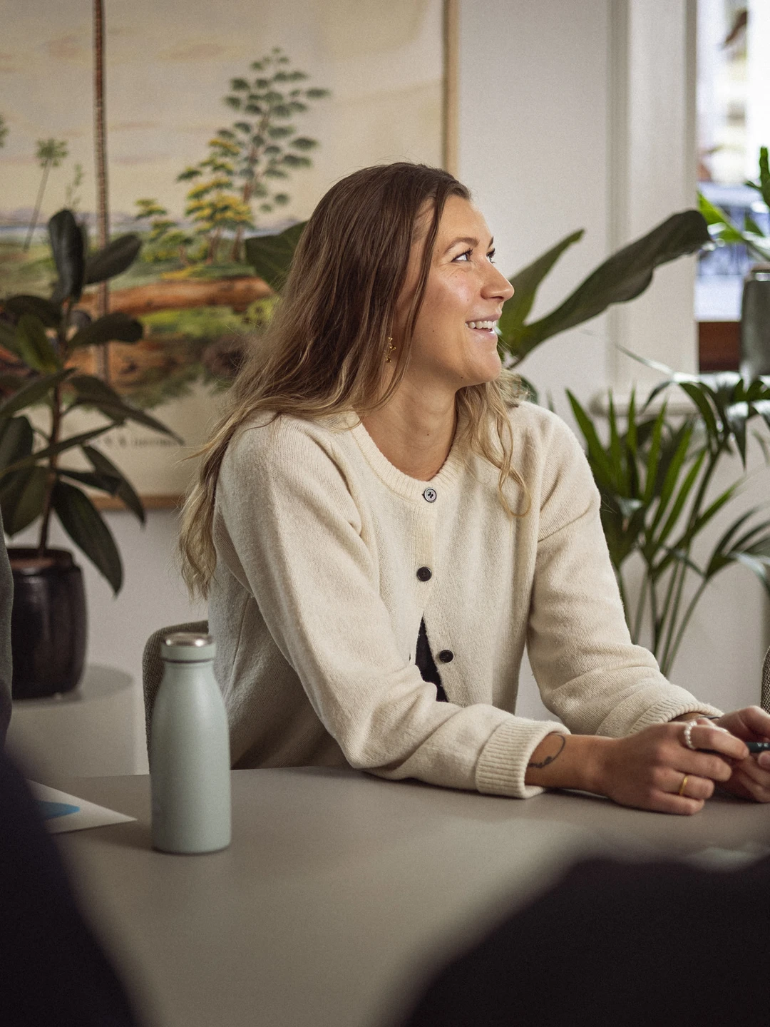 En gruppe mennesker deltager i en arbejdsbeskrivelse, hvor en smilende kvinde i en lys trøje sidder ved et bord med planter i baggrunden.