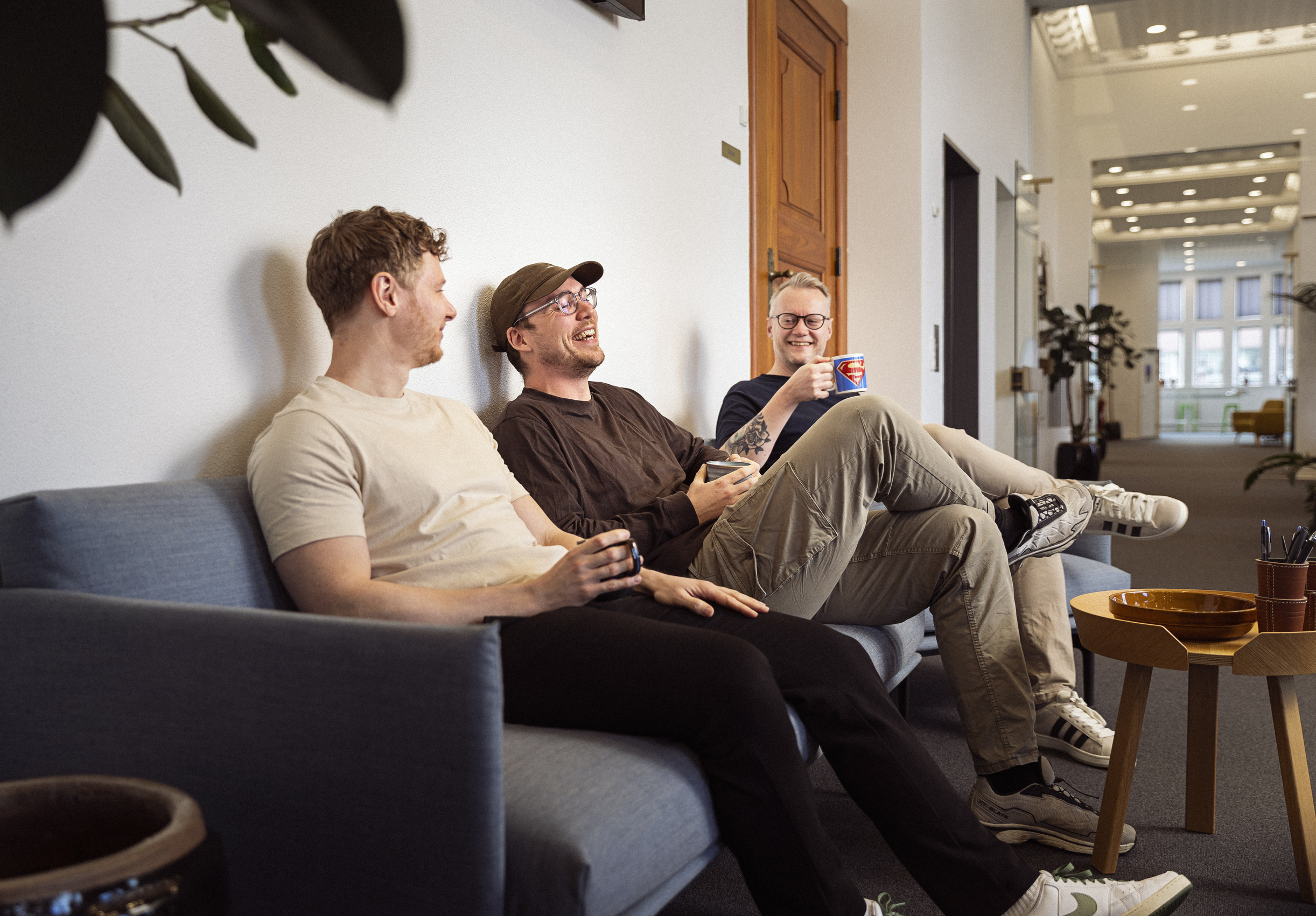 Tre mænd sidder på en sofa i et lyst lokale, griner og drikker kaffe, med planter og en træbord i baggrunden.