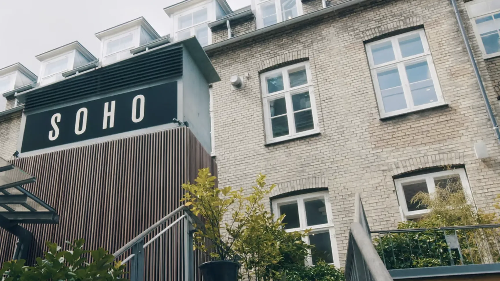 Billede af bygningen "SOHO" med en moderne facade, trapper, planter og vinduer, under en skyet himmel.