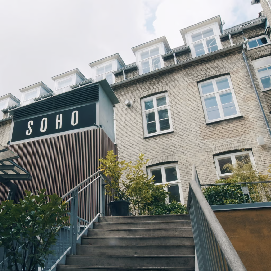 Billede af bygningen "SOHO" med en moderne facade, trapper, planter og vinduer, under en skyet himmel.