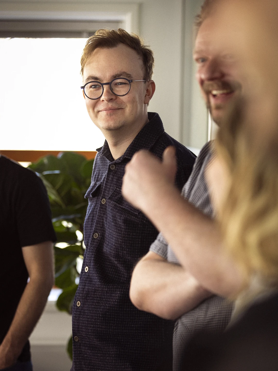 En gruppe mennesker i en lys kontorindstilling, hvor en smilende mand med briller og kort, lys hår står i fokus, mens andre personer delvist ses i baggrunden. Der er grønne planter i rummet, hvilket skaber en indbydende atmosfære.