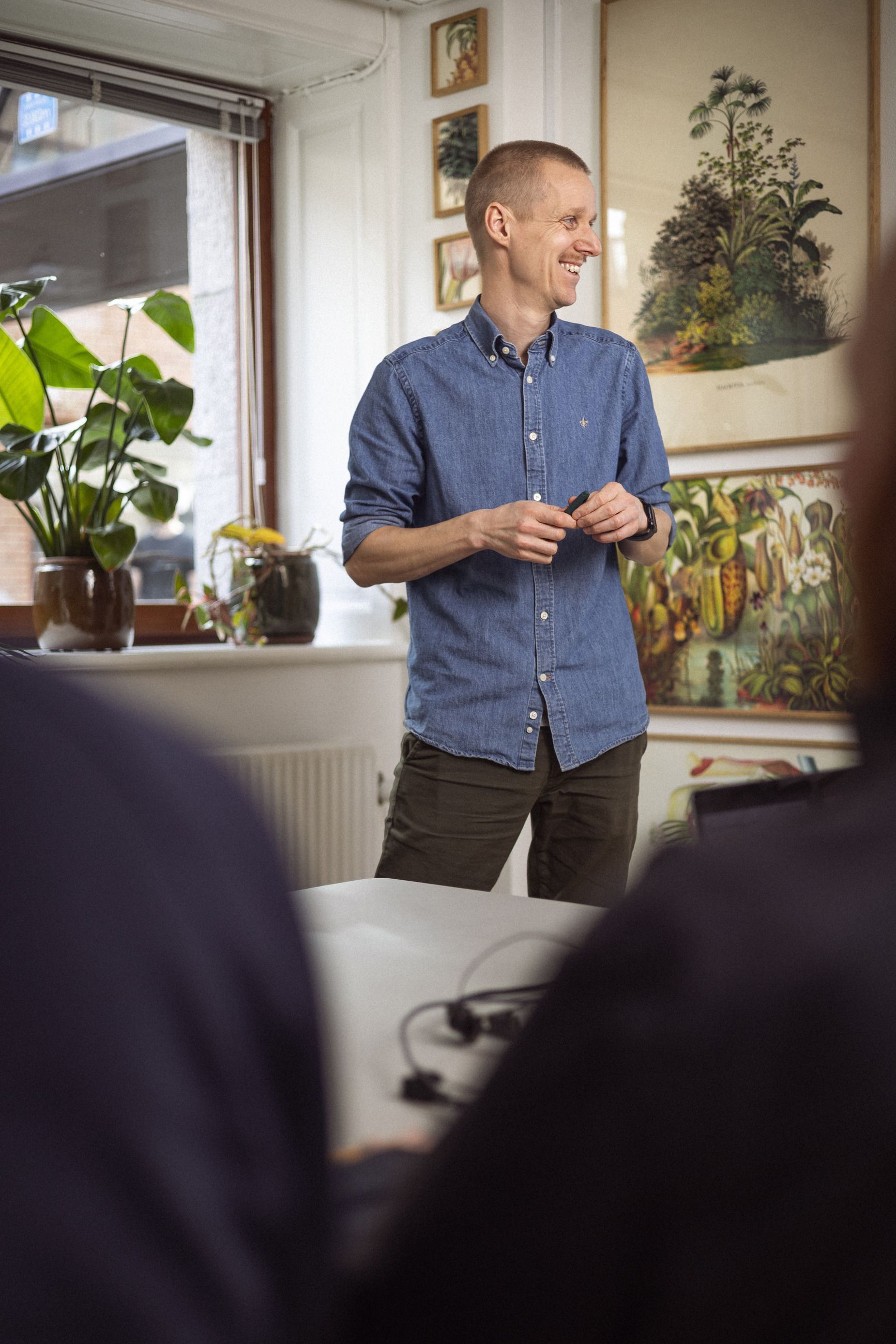 En smilende mand i en blå skjorte præsenterer foran en gruppe mennesker i et lyst rum med planter og farverig vægkunst.