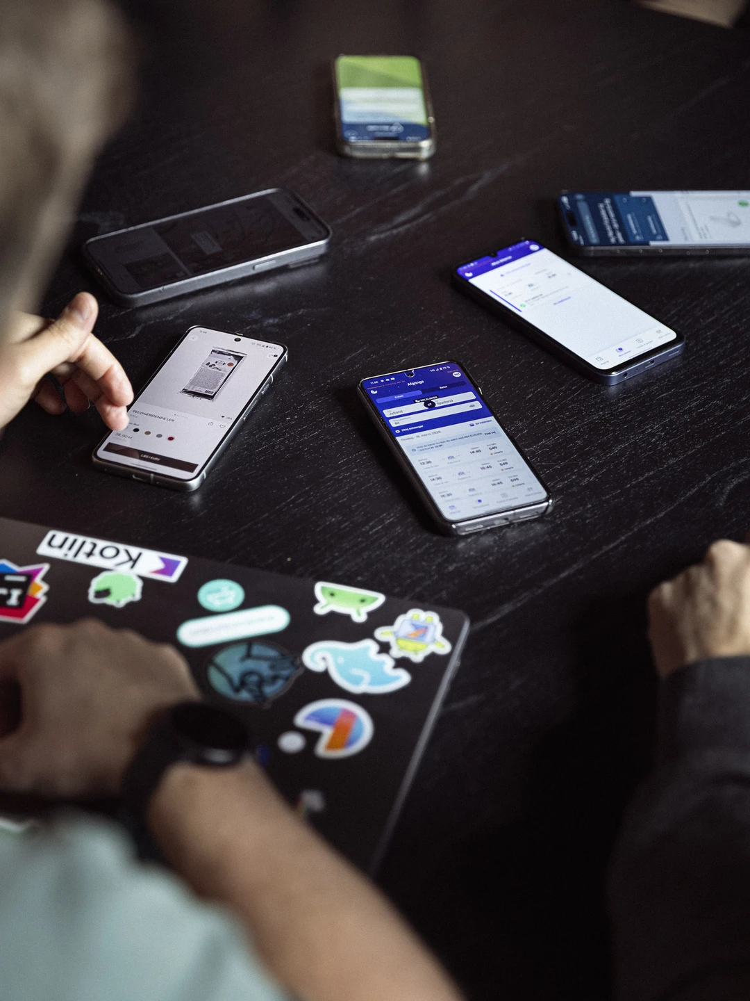 En gruppe smartphones på et bord, med fokus på skærmbilleder og forskellige apps, samt en hånd, der interagerer med en laptop med klistermærker.
