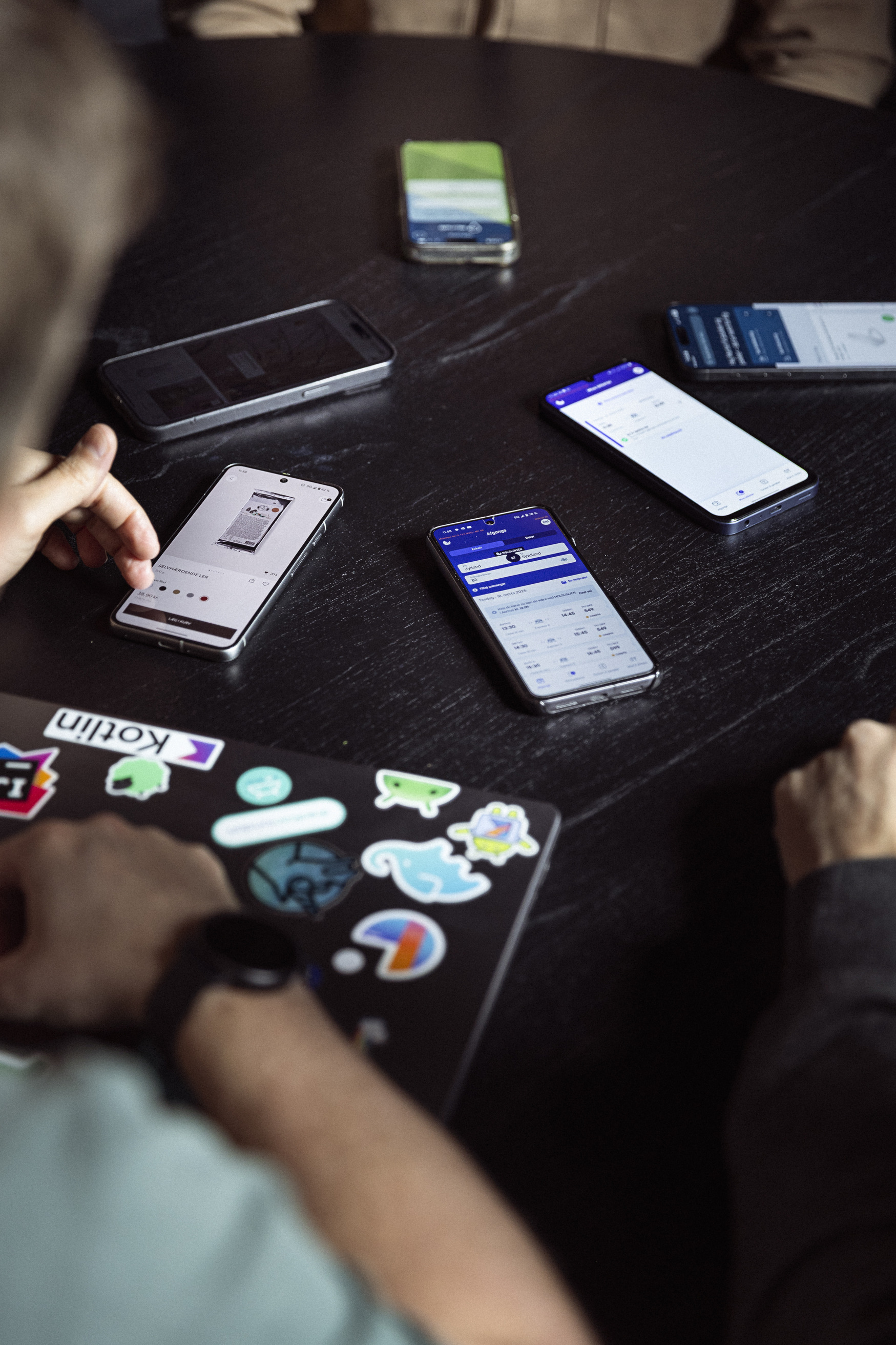 En gruppe smartphones på et bord, med fokus på skærmbilleder og forskellige apps, samt en hånd, der interagerer med en laptop med klistermærker.