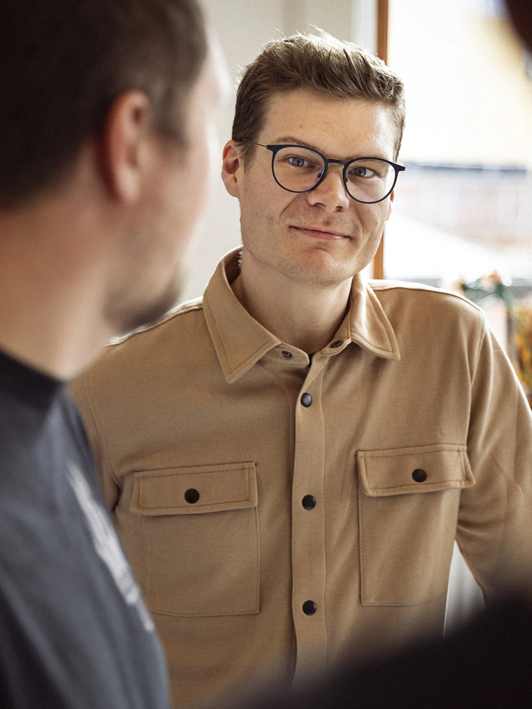 Billede af en ung mand med briller og et venligt smil, iført en beige skjorte med lommer, der står i et lyst og indbydende rum, mens han interagerer med andre personer i en uformel samtale.